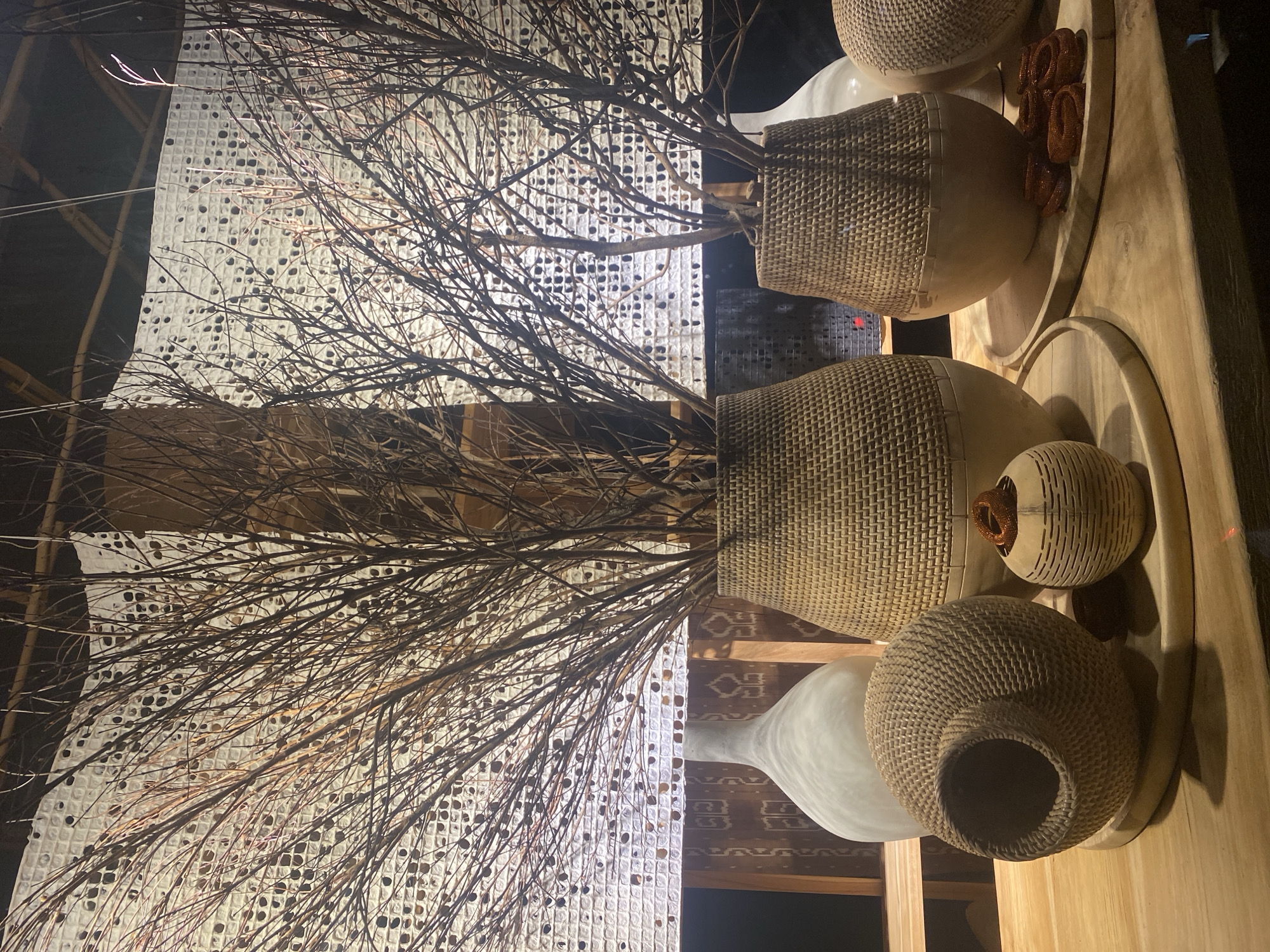 A still life arrangement of decorative objects is displayed on a wooden surface. In the background, a white, textured screen with circular cutouts is illuminated from behind, creating a patterned light. Various vases and decorative vessels are placed on wooden trays.
Dominating the scene are tall, dry branches with numerous small twigs, fanning out from within woven vases. One large, light brown woven vase with a smooth, rounded bottom is centrally located, holding a dense cluster of these branches. To its left, a tall, slender off-white ceramic vase with a rounded body and narrow neck stands. To the right of the large woven vase, a smaller, similar woven vase holds a sparser collection of the dry branches. Further to the right, another large woven object, possibly a basket or vase, is partially visible.
In the foreground, several smaller decorative items are arranged. A spherical, textured woven object with a large opening is positioned to the left. Beside it, a small, cylindrical woven vessel with decorative horizontal grooves contains a coiled, reddish-brown decorative element. In front of the smaller woven vase, a cluster of small, dark reddish-brown coiled decorative objects rests on a wooden tray.
The overall aesthetic suggests a natural and earthy ambiance, possibly a retail display or a home decor setting. The lighting is warm and somewhat dim, highlighting the textures of the objects. The image appears to be taken indoors, with the background suggesting a ceiling structure and possibly a wall.
Without additional information or contextual clues within the image itself, it is impossible to definitively determine the specific location within Denpasar, Indonesia. The scene depicts decorative items, not specific landmarks or identifiable public spaces that would allow for precise geographical localization.
