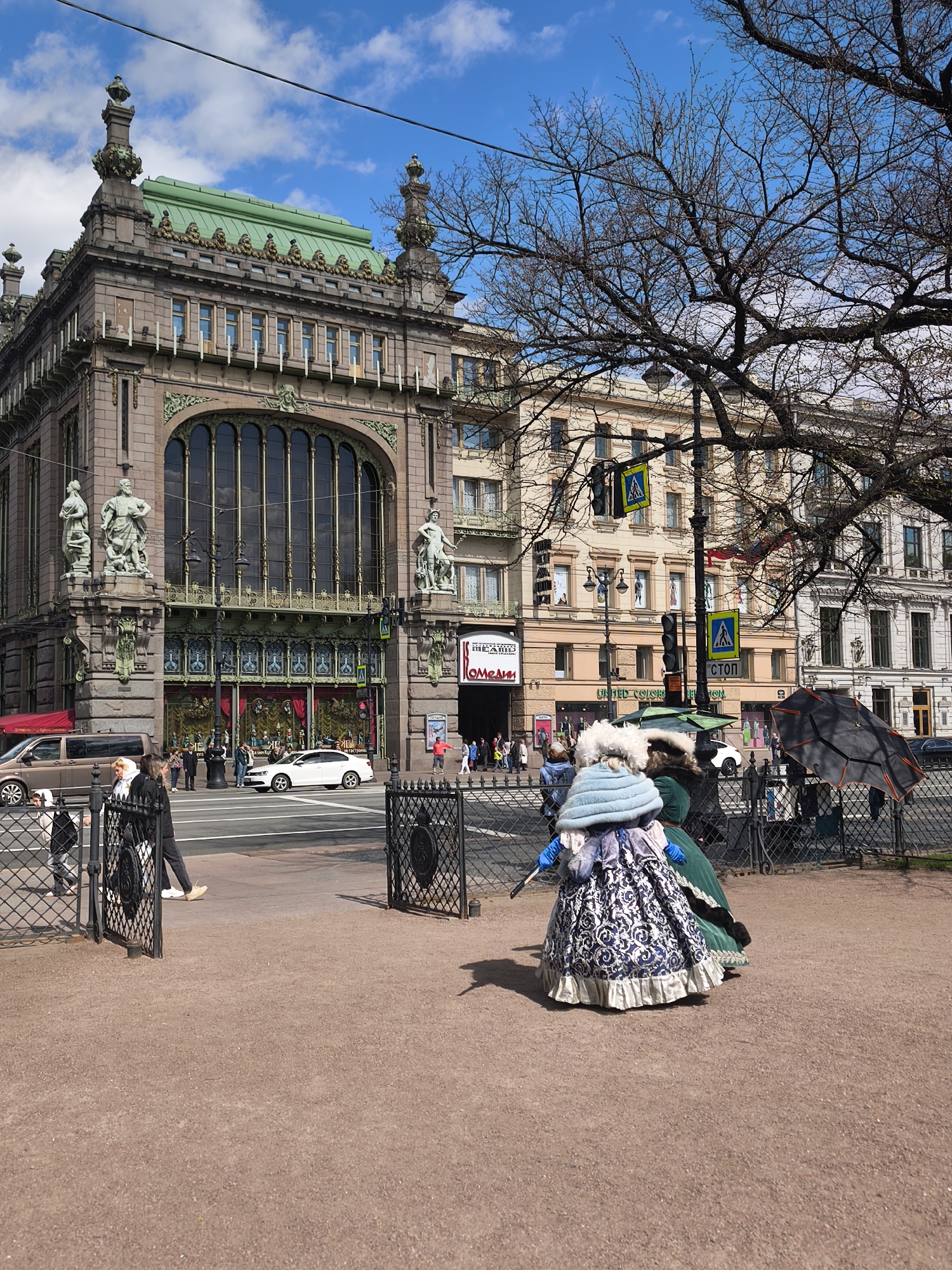 The image depicts a street scene in Saint Petersburg, Russia, featuring historical architecture and modern elements. In the foreground, there is an open wrought-iron fence leading onto a gravel path. Two individuals dressed in ornate, historical costumes stand on the gravel path. One costume is predominantly blue and white with a large, layered hat, while the other is green. Behind the fence, pedestrian traffic crosses the street. There are vehicles including a dark-colored minivan and a white sedan. In the background, architectural details of historical buildings are visible, including one building with a green roof and numerous windows. A pedestrian crosswalk sign and a traffic light are present. The sky is partly cloudy, and the branches of a large tree are visible on the right side of the image. A building features signage in both English and Russian. The scene appears to be a mix of historical reenactment and contemporary urban life.