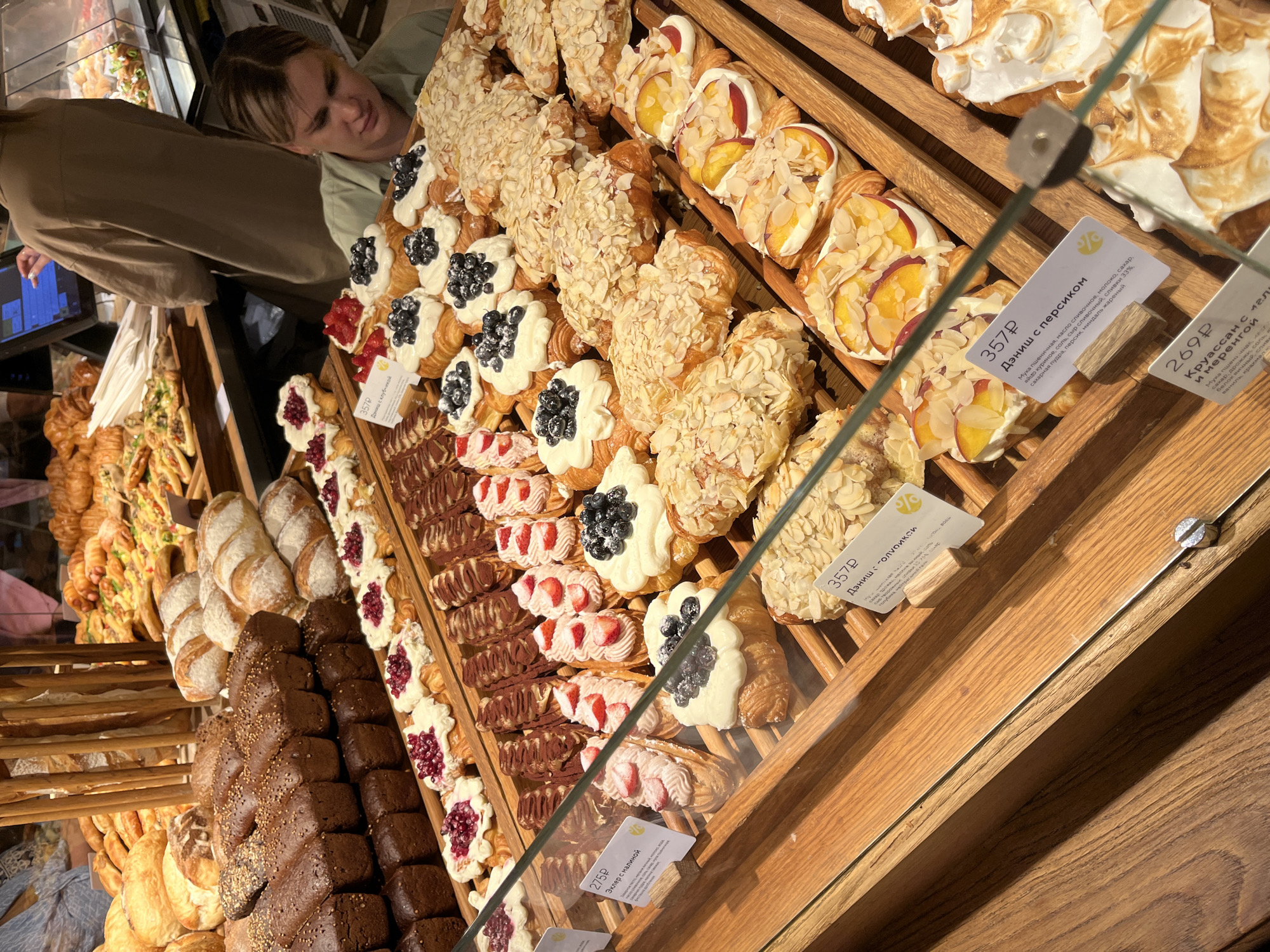 The image displays a bakery display filled with a variety of pastries, breads, and other baked goods. In the foreground, a glass barrier separates the viewer from the display. Several types of croissants are visible, some topped with blueberries and cream, others coated in sliced almonds. There are also pastries with fruit toppings, possibly strawberries or raspberries, and some that appear to be chocolate-dipped. Loaves of dark bread are stacked in the background, alongside other types of bread and possibly pies or tarts. A person with light-colored hair is partially visible in the upper right corner, wearing a light-colored top, looking down towards the display. In the background, another person wearing a brown garment is interacting with a touchscreen device. The overall environment suggests a retail bakery setting, with wooden display shelves and visible pricing labels in Russian currency. The scene is well-lit, highlighting the textures and colors of the baked goods. The presence of Russian pricing labels and the overall aesthetic are consistent with a bakery in Svetlogorsk, Russia.