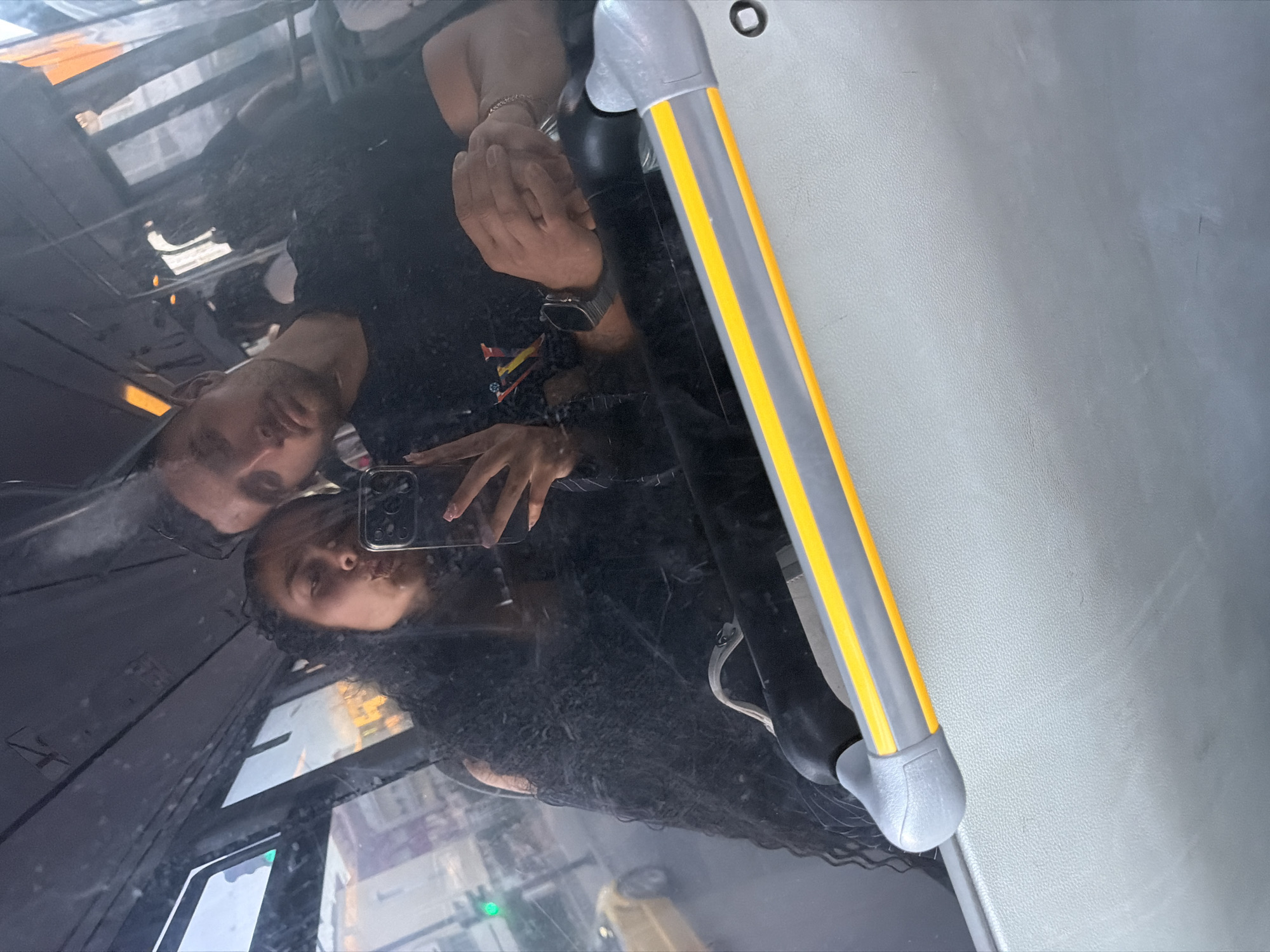 A young couple takes a selfie in a bus. The woman in the foreground puckers her lips for the camera, holding her phone in her right hand. She has long, dark, curly hair and is wearing a black, fuzzy jacket. Beside her, a man leans his head against hers, his eyes partially closed. He has short dark hair and is wearing a black t-shirt. They appear to be in motion, as the reflection in the bus window shows a blurred urban landscape with buildings and a yellow car. The bus interior features a gray seat and a handrail with yellow and gray stripes. The photo was taken in Nesebar, Bulgaria.