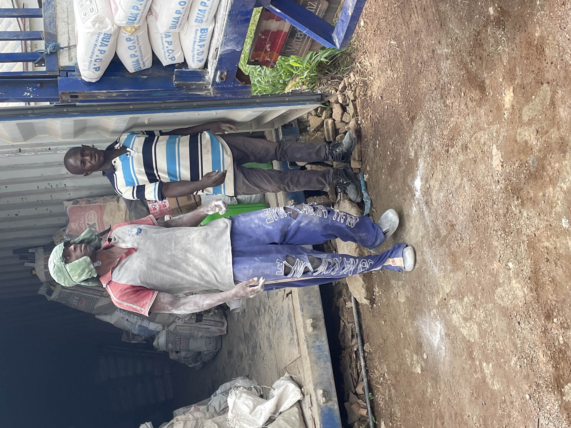 In Enugu, Nigeria, two men are standing in front of an open shipping container filled with bags of cement. The man on the left is wearing a red and grey shirt, ripped blue trousers, and a green head covering. The man on the right is wearing a blue, white, and black striped polo shirt and dark trousers. The ground is dirt and gravel, with some scattered debris. The shipping container appears to be parked in an outdoor area, possibly a warehouse or construction site. The bags of cement in the container are labeled "BUA P.O.P. Special Moulding Plaster."