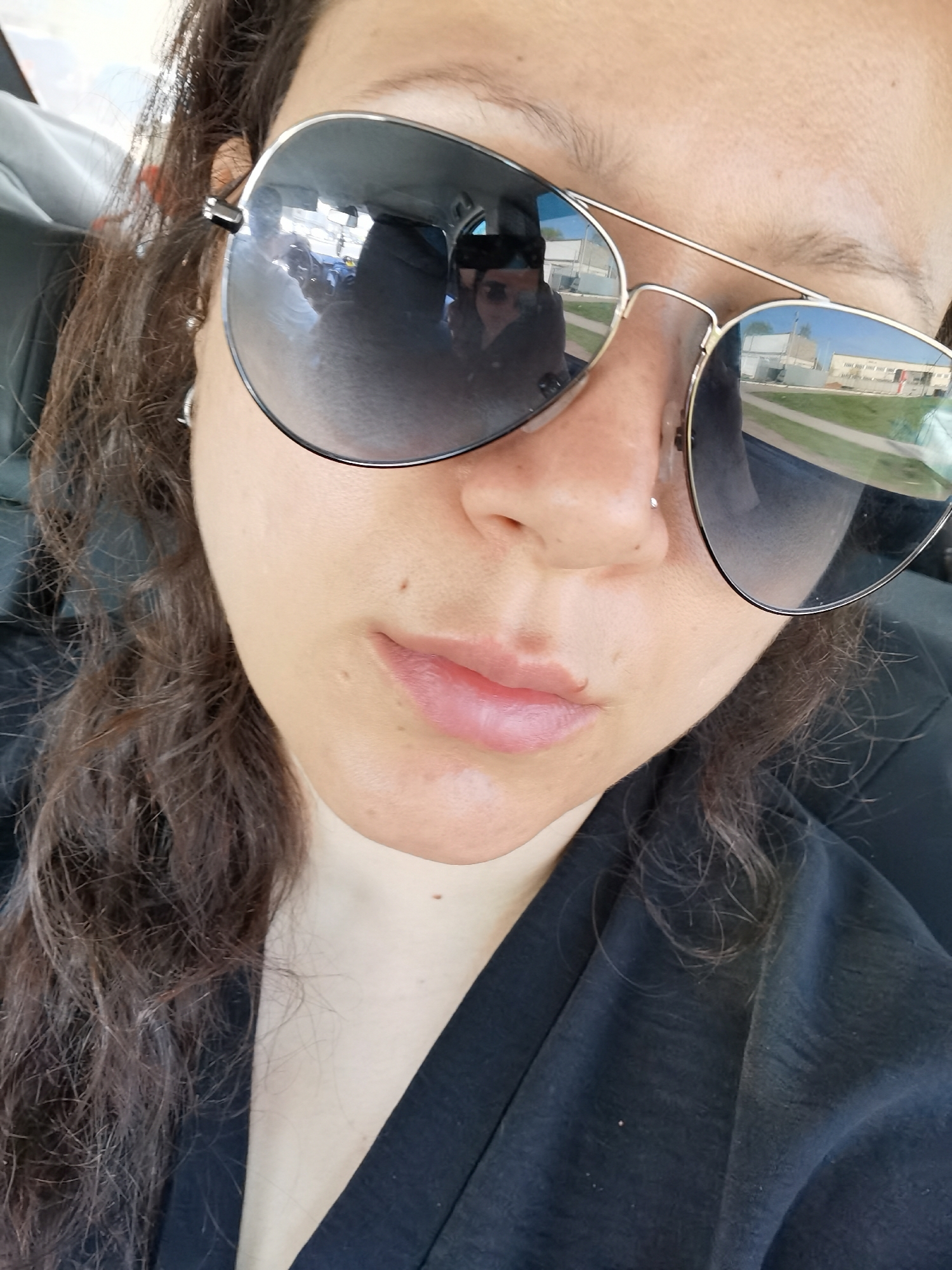 A close-up selfie of a woman in a car, wearing aviator sunglasses. The reflection in the sunglasses shows a building and green grass. The building is the Филиал ГБПОУ “Чайковский индустриальный колледж” in the city of Chaikovsky, Russia. The woman has dark curly hair, light skin, and is wearing a black shirt. She has a piercing in her nose and earrings. The car interior and other occupants can be seen blurred in the sunglasses' reflection.