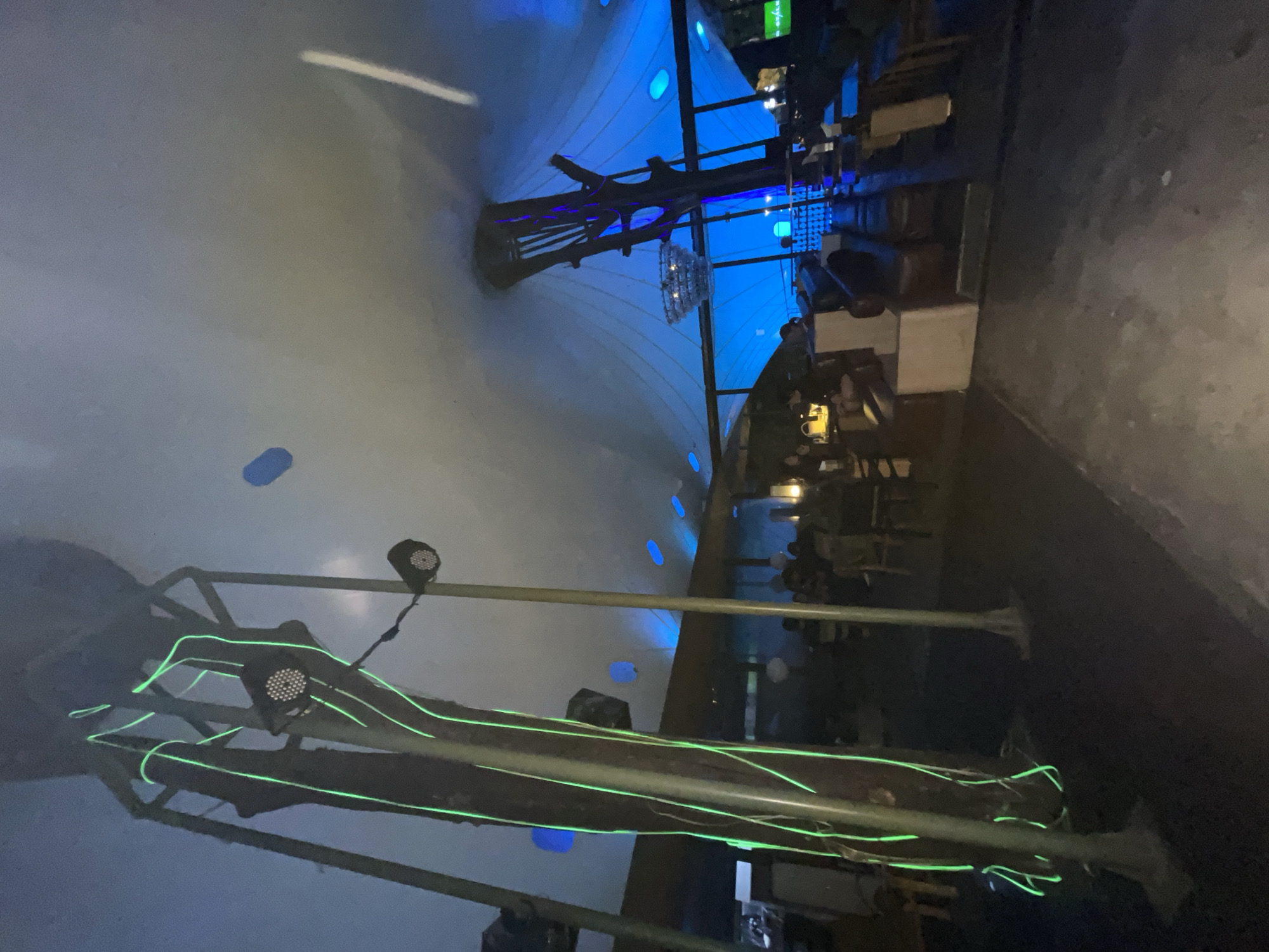 The image depicts the interior of a dimly lit establishment, possibly a restaurant or bar, in South Jakarta, Indonesia. The most prominent feature is a large, tent-like structure overhead, illuminated with blue and white light. Decorative light fixtures are suspended from the ceiling structure. There are tables and chairs arranged within the space, suggesting a dining area. Additional decorative lighting, in green, is visible on what appears to be a tree branch or similar architectural element. The overall ambiance is modern and atmospheric.