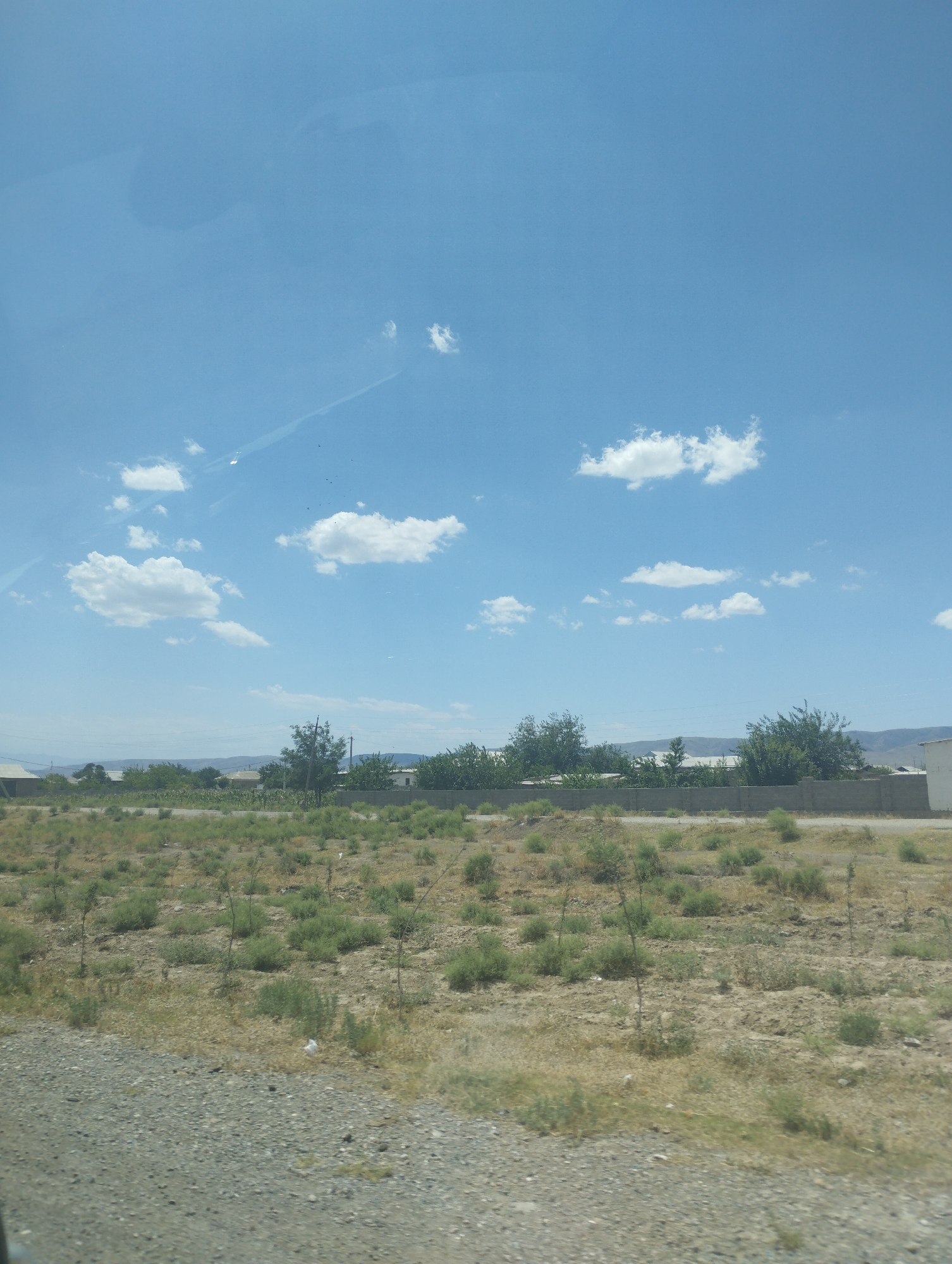 This is a full-frame, eye-level, medium shot of a natural outdoor environment under a bright blue sky. The upper portion of the image is dominated by the sky, which features scattered white cumulus clouds. There are faint, linear cloud formations or contrails visible, suggesting aerial activity.
The middle ground of the image displays a landscape with low-lying vegetation and scattered trees. A concrete wall runs horizontally across this area, separating the foreground from the background. Beyond the wall, several buildings with light-colored roofs are visible, interspersed with more trees. The overall impression is that of a rural or semi-urban fringe in a dry climate.
The foreground consists of a gravelly ground surface, with patches of dry grass and small green bushes. The textures of the gravel and the sparse vegetation suggest a dry, possibly arid, environment. The lighting indicates it is daytime, with the sun likely high in the sky, casting a bright, even light.
While the image is identified as being in Egizbulak, Uzbekistan, there are no specific landmarks or features in the photograph that can definitively confirm this location without additional context. The scene is devoid of any people, animals, or discernible actions, presenting a static environmental snapshot.