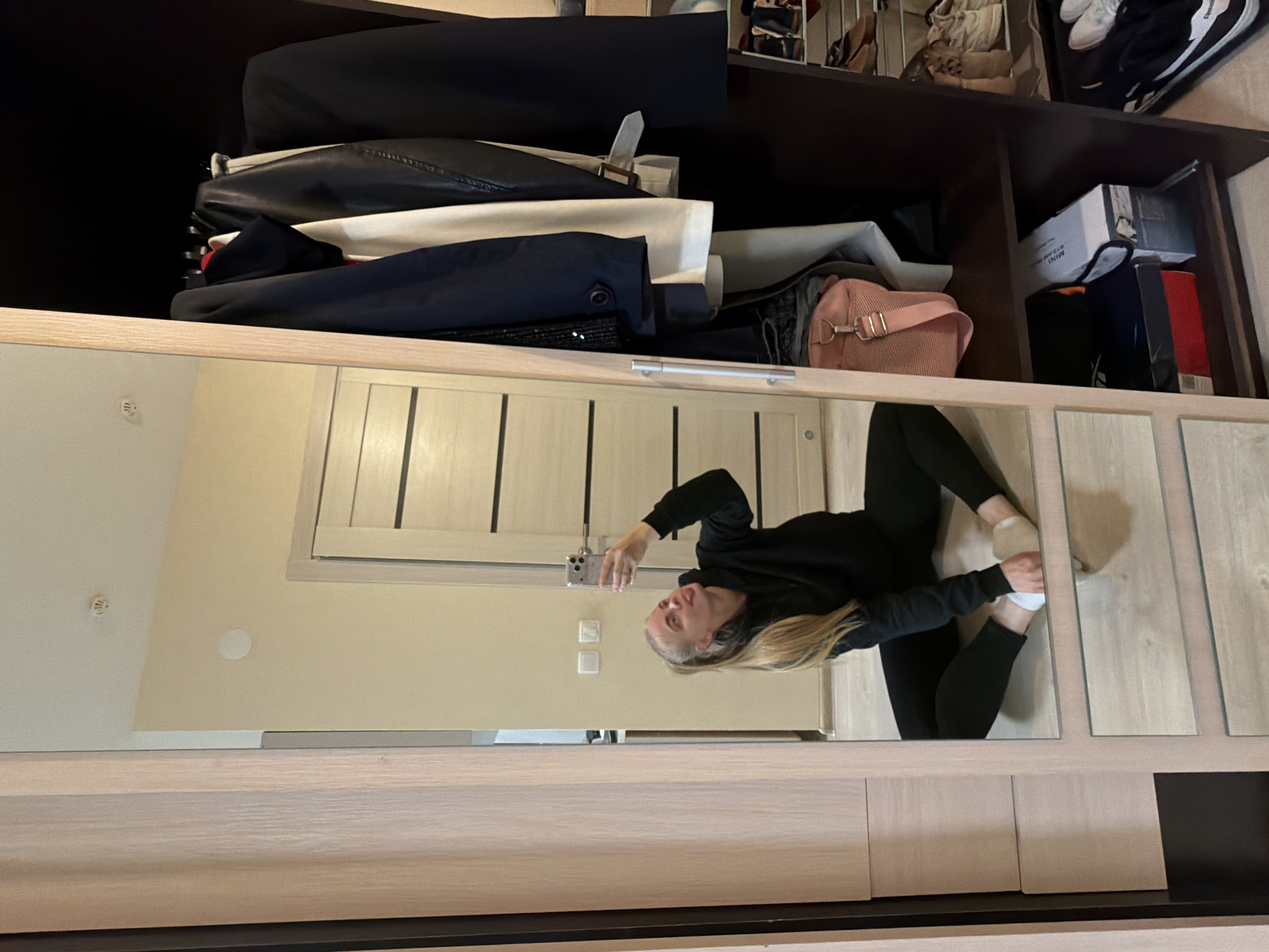 A woman is taking a photo of herself in a full-length mirrored closet in Izhevsk, Russia. She is wearing black leggings and a black hoodie and white socks. The closet contains clothing, boxes, and shoes. The interior of the closet and the surrounding walls are beige and wooden. The mirror is paneled.
