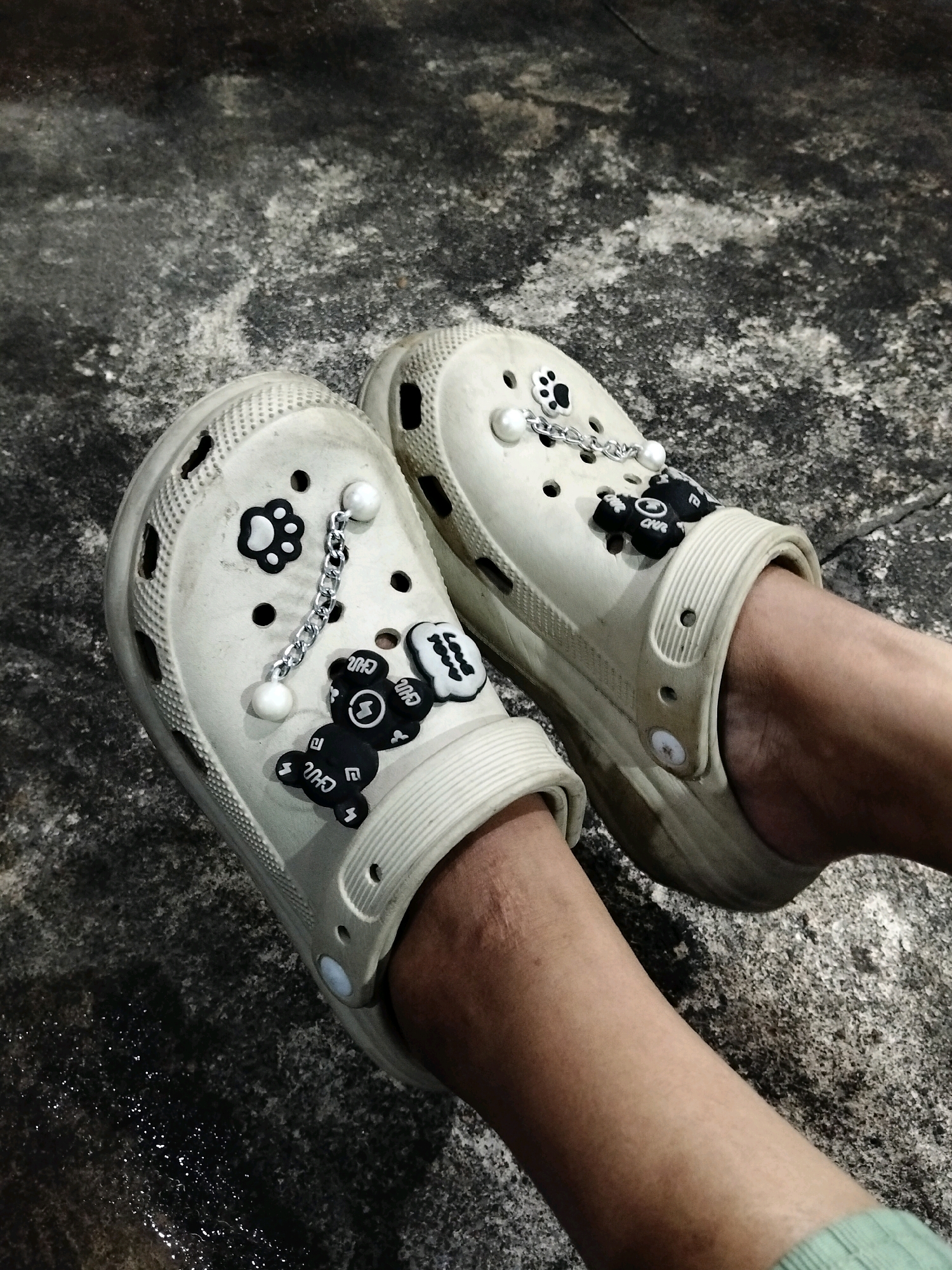 The image shows a close-up view of a pair of beige Crocs adorned with black charms shaped like cartoon bears and white pearls, attached by silver chains. The shoes are on a person's feet, standing on a wet, grey, mottled surface that appears to be concrete, possibly a sidewalk or road. The lighting suggests an outdoor setting, possibly at night or in a dimly lit area. The background is dark and out of focus, with some visible texture on the ground. The location is Arang Limbung, Indonesia.