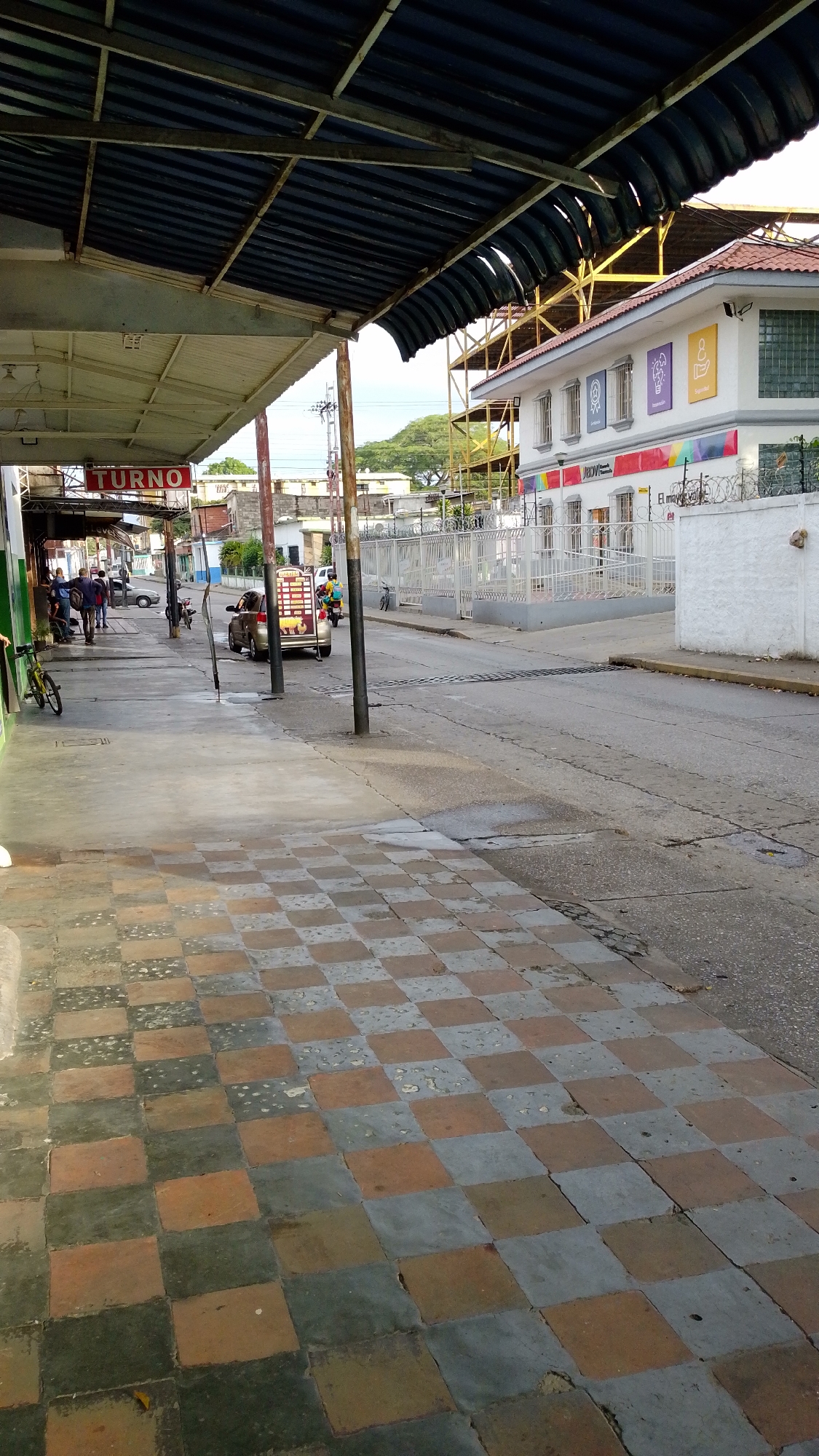 The image shows a street scene in Tinaquillo, Venezuela. The foreground displays a checkered tiled sidewalk, alternating between light brown and gray. The street is made of cracked asphalt, and a white building is visible across the street with multi-colored square emblems visible near the top of the building, along with a barbed wire fence. A few people are on the sidewalk under a covered awning, and a car and a motorized cart are in the street. Electrical poles are also visible along the street.
