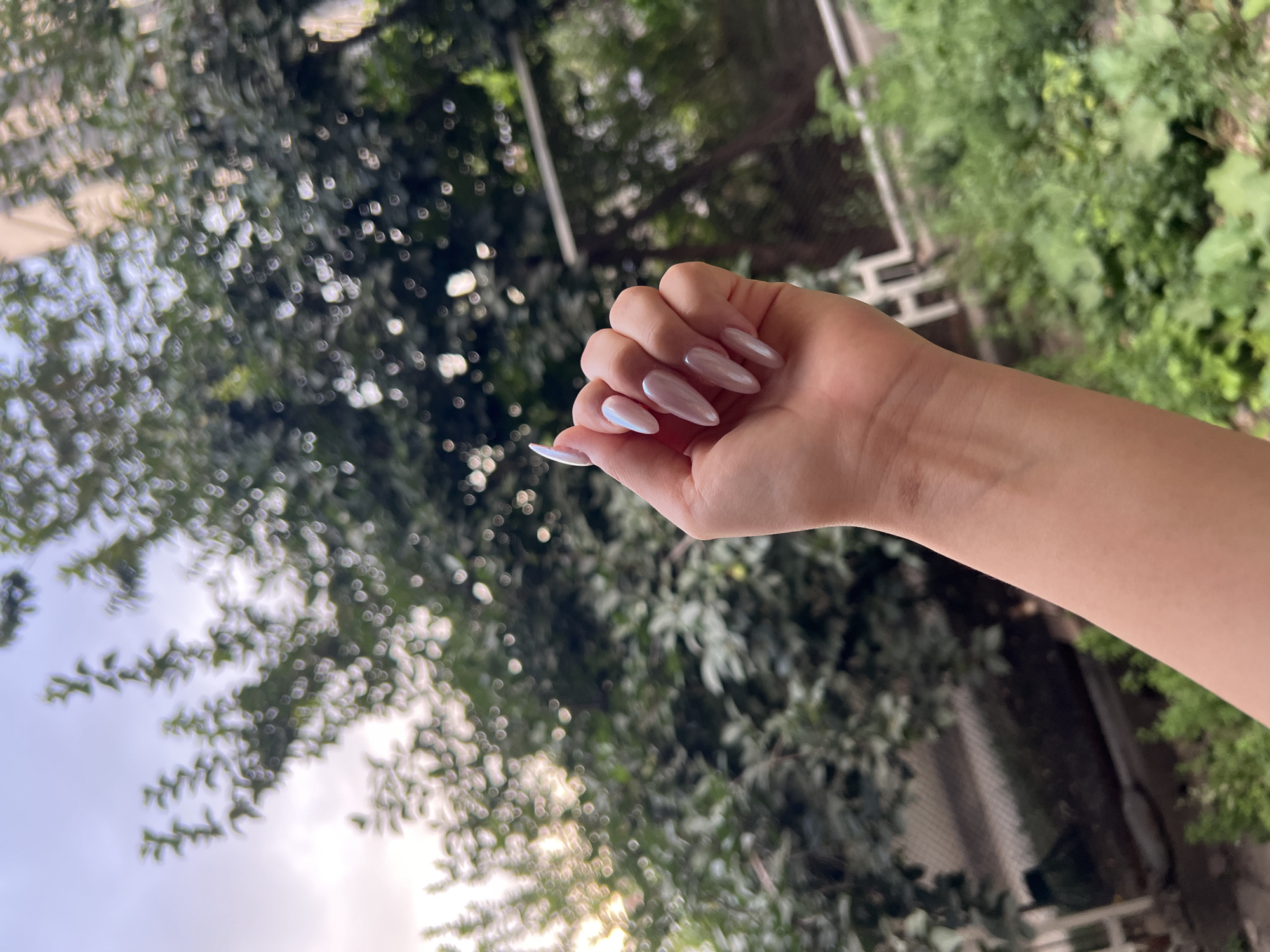 A hand with long, almond-shaped fingernails painted a pearlescent, light pink color is held up in the foreground. The hand is angled to show the fingers splayed slightly. Behind the hand, the background is a softly blurred scene of lush green trees and vegetation. The sky is visible above the trees, with soft clouds and hints of sunlight. The scene appears to be outdoors, possibly in a garden or park. The image does not contain any identifiable landmarks or elements that specifically place it in Osh City, Kyrgyzstan.