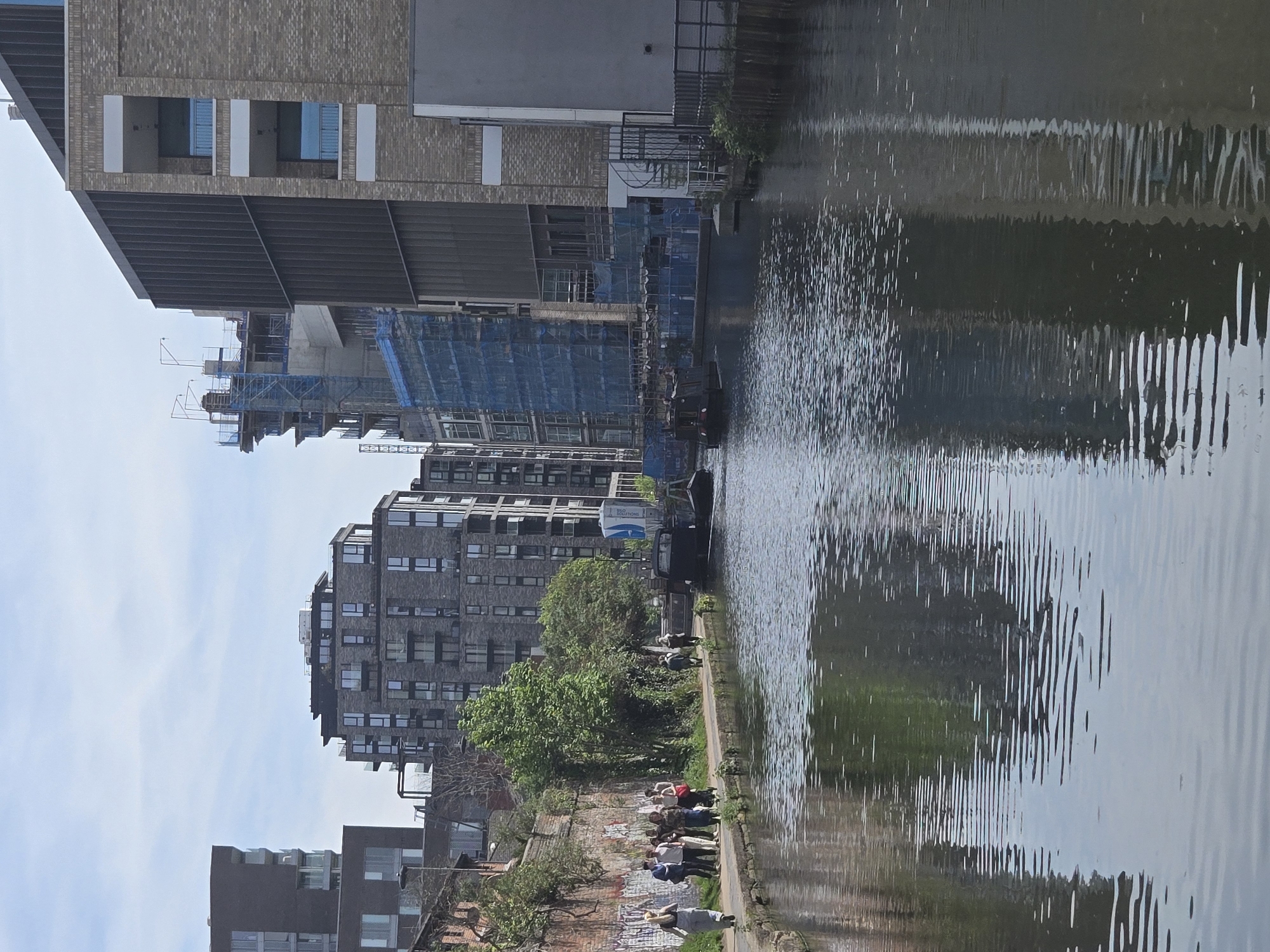 The image shows a canal lined with buildings in what appears to be a city. Several buildings are visible, including some under construction with scaffolding and blue netting. The canal's water reflects the buildings and the sky. A narrow walkway runs along the canal's edge, with people visible on it. In the foreground, individuals stand and sit along the walkway, some appearing to be interacting. The canal has a barge in it. The buildings are modern, varying in height and design, some with multiple windows. The location is in London, United Kingdom.