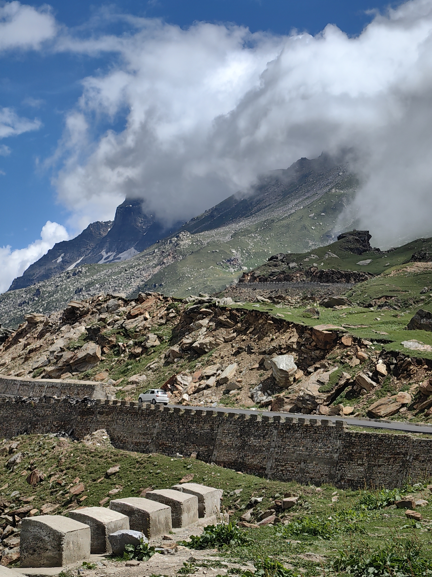 The image captures a mountainous landscape with a road winding along the side of a steep, rocky slope. A white car is seen driving on the road, which is bordered by a stone wall. The mountains in the background are partially obscured by clouds, with patches of green vegetation and snow visible on their slopes. The foreground shows a rugged terrain with scattered rocks and some concrete structures. The scene is likely located in or around Gramphu, India, a region known for its high-altitude roads and scenic mountain views. The sky is partly cloudy with blue patches.