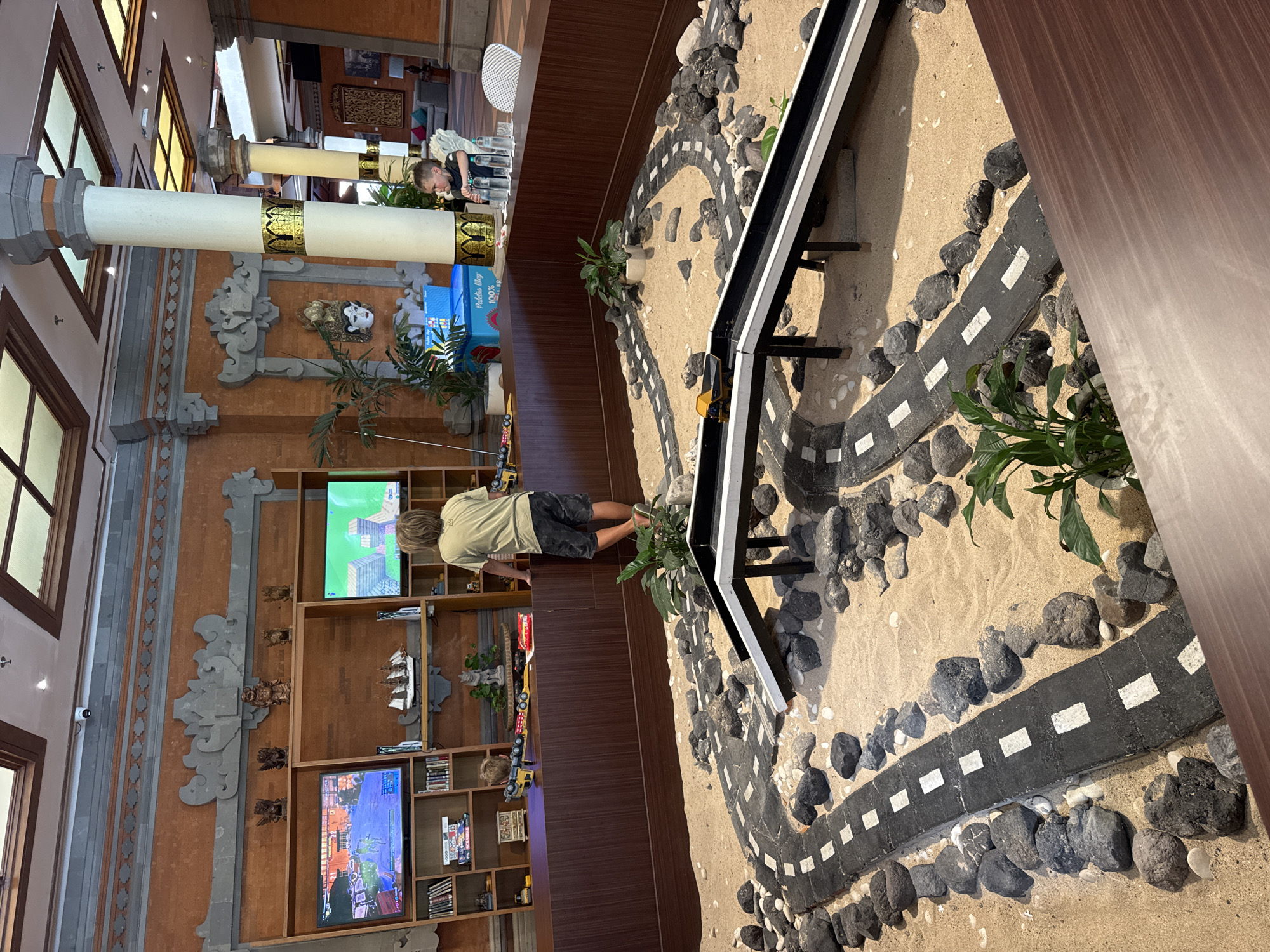 This image captures an indoor recreational area, likely within a resort or hotel, in Kuta Selatan, Indonesia. The scene is dominated by a children's play space in the foreground and a media/activity wall in the background, set within an environment featuring traditional Balinese architectural elements. In the immediate foreground, a rectangular sandpit is visible, filled with light-colored sand. Within the sandpit, a miniature road system, constructed from dark gray segments with white dashed lines, winds through the sand. Numerous dark gray and black rocks are strategically placed along the edges of the roads and scattered throughout the sand. Two elevated black and white ramps, supported by black posts, form part of the road network, with a small yellow toy dump truck positioned on one of the ramps. Several small, potted green plants are interspersed within the sandpit. Separating the sandpit from the background is a dark brown wooden counter. On the left side of this counter, a yellow and black toy construction vehicle and a smaller red toy car are present. A young boy, wearing a light-colored t-shirt and dark shorts, stands on the counter with his back partially to the camera, facing two television screens mounted on a shelving unit. To the right, another young boy, dressed in a dark t-shirt, is seated in a white high chair at the counter. He holds a small, dark object in his hands, and several clear glasses filled with water are arranged in front of him on the counter. Behind the counter is a large wooden shelving unit integrated into the wall. This unit houses two large flat-screen televisions. The television on the left displays a dynamic video game scene, possibly a third-person shooter or adventure game, with characters and on-screen interface elements visible. The television on the right displays a block-building video game. Below the televisions, the shelves are populated with books, some stacked and some upright, and various board games, including "Scrabble." Decorative objects, such as several carved wooden or stone figurines and a model sailboat, are also displayed on the shelves. A small white statue is visible in a planter beneath the model sailboat. A large blue box with "Paletas Way" printed on it is positioned behind the counter, near a pillar. The background features terracotta-orange walls with intricate gray stone decorative friezes and panels, indicative of Balinese craftsmanship. Two prominent, tall, round pillars, white/cream-colored with decorative gold and