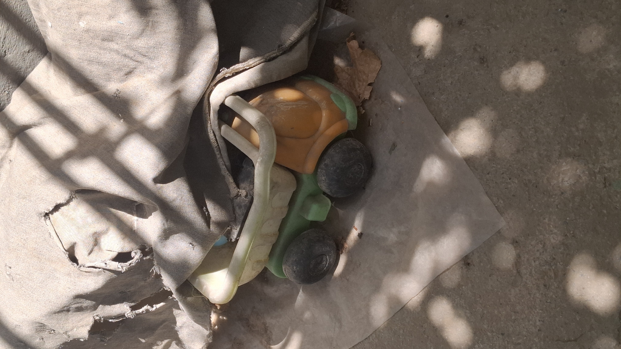 An outdoor scene in Шұбарсу, Kazakhstan depicts a partially covered toy dump truck. The dump truck, predominantly orange and green with black wheels, is situated on a concrete surface, partially obscured by a torn, light-colored fabric draped over it. The fabric shows signs of wear and tear, with visible holes and fraying. Sunlight casts dappled shadows across the scene, suggesting it is daytime. A thin, translucent sheet of material, possibly plastic, lies beneath the toy. A dry leaf is visible near the rear of the truck. The surrounding environment appears to be an outdoor area, possibly a yard or alleyway, with the concrete ground showing some debris and discoloration.
