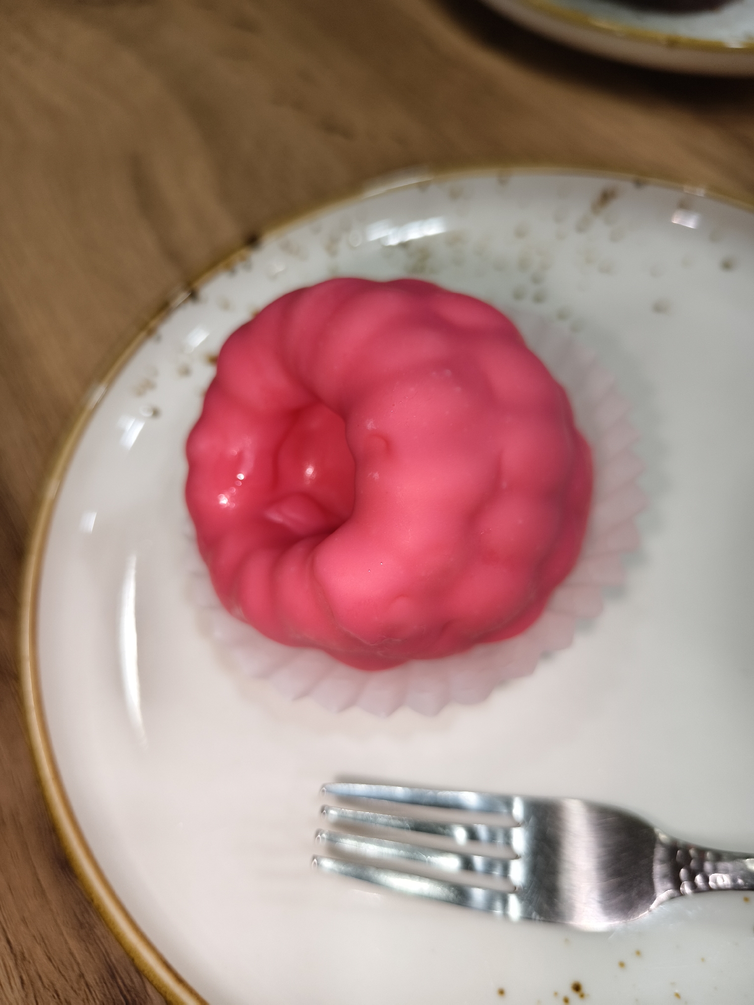 A close-up, overhead shot shows a pink frosted cake or pastry on a white speckled plate with a gold rim. The pastry is nestled in a white paper liner and has a rounded, irregular shape with visible frosting textures. A silver fork with three tines rests on the plate to the right of the pastry. The background is a wooden surface. The scene appears to be indoors, possibly a cafe or home setting, in Unknown, Unknown. There are no people or specific city landmarks visible.
