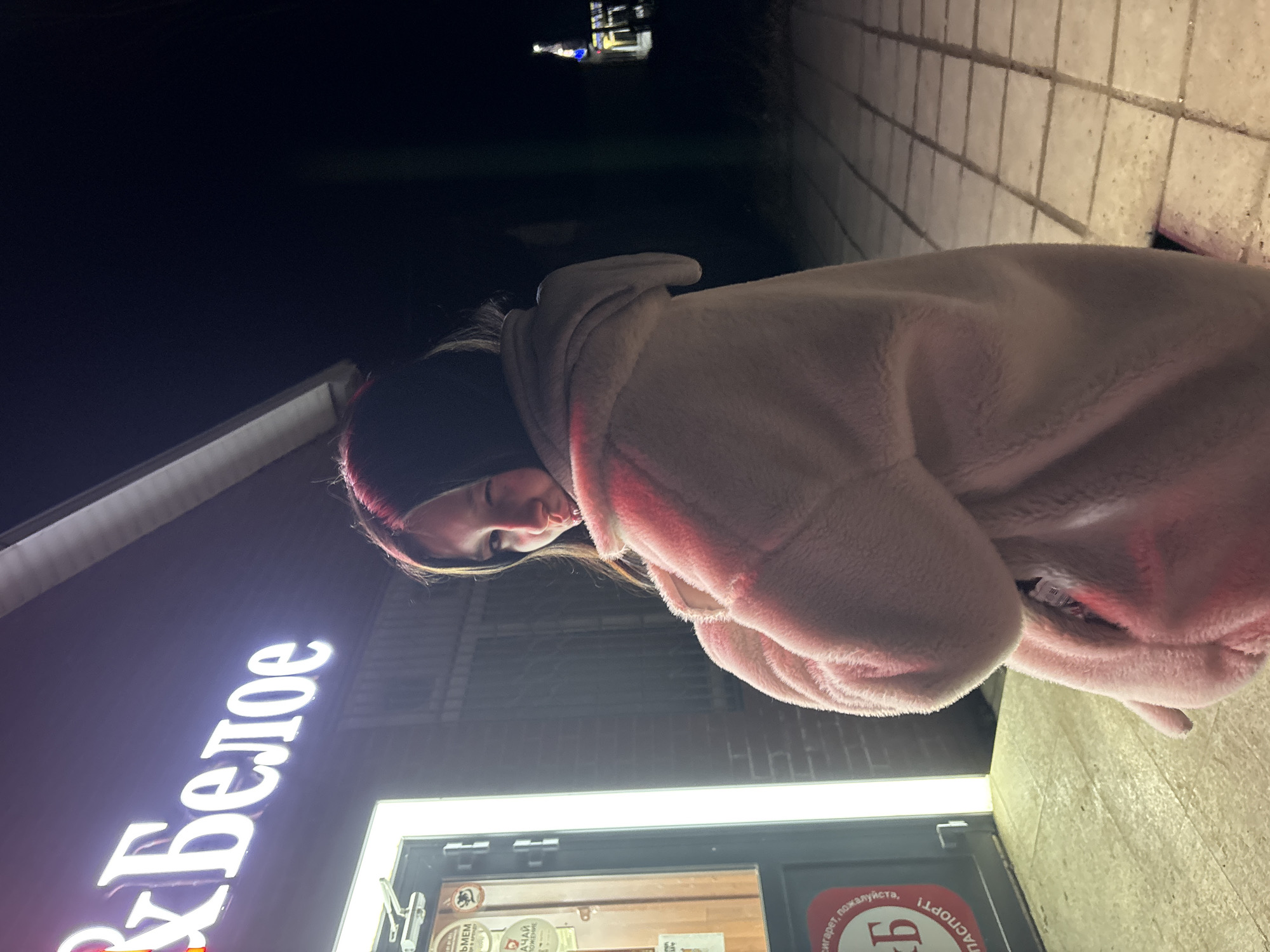 A person stands next to the brightly lit storefront of a "БХ Белое" store at night in the city of Фоки, Russia. The individual, seen from a slightly tilted angle, is wearing a dark head covering and a light pink, plush hooded jacket. The store's signage is predominantly white, and the entrance features bright fluorescent lighting above the door. Outside the store, the sidewalk consists of light-colored tiles. The background features a dark night sky with some lights visible in the distance.