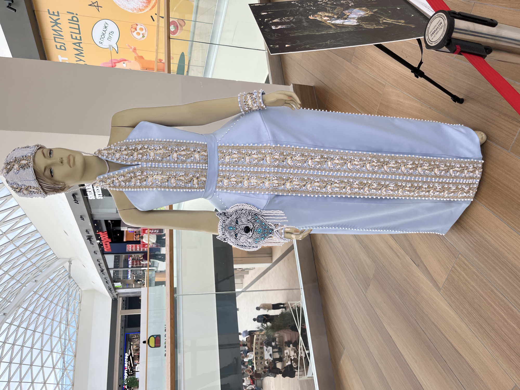 The image shows a mannequin dressed in a light blue, floor-length dress adorned with intricate beadwork and pearl embellishments. The dress features a halter neckline and a fitted bodice, with decorative trim running vertically down the front and along the hem. The mannequin is also wearing a matching headpiece and a wide bracelet on its right wrist, decorated with a wolf's head design. A similar, smaller bracelet is on its left wrist. The mannequin is displayed indoors, likely in a public space given the surrounding architecture and signage. In the background, there is a large glass ceiling with a metal grid structure, indicating a modern building. Further back, there are shops with visible signage, including "maxxpert" and "SNEAKERS STREET". The floor is made of light-colored wooden planks. To the right of the mannequin, a framed artwork is displayed on a tripod easel, with a red velvet rope barrier in front of it. A sign on the rope barrier contains text in Russian. The overall environment appears to be a shopping mall or an exhibition space. The text on the orange advertisement in the background appears to be in Russian, and based on the visual cues and context, this scene is likely located in the city of Perm, Russia, specifically within the "Planeta" shopping center, known for its distinctive glass ceiling and atrium.