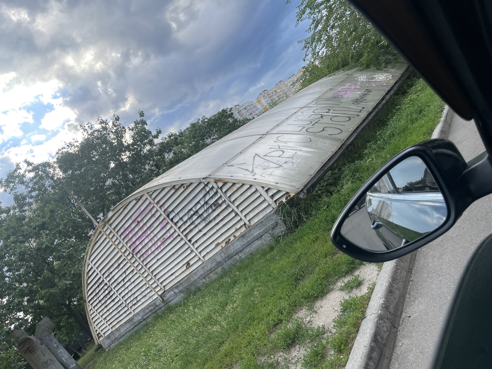A low-angle, wide shot captures an overcast day in Kyiv, Ukraine. The foreground is dominated by a car's wing mirror, reflecting the sky and buildings. To the left, a long, curved structure, possibly an old bus stop or shelter, is covered in graffiti. This structure is made of a translucent, corrugated material supported by a metal frame with horizontal slats. Pink and black spray paint tags mar its surface.
Behind the shelter, lush green trees line the background, with apartment buildings visible in the distance. The sky is a mix of heavy, dark clouds and brighter patches, suggesting intermittent sunlight. The ground is covered in grass and dirt, with a paved road and curb on the right side of the frame. Two large metal pipes are visible in the lower left corner, also marked with graffiti. The overall impression is one of an urban fringe environment on a cloudy day.