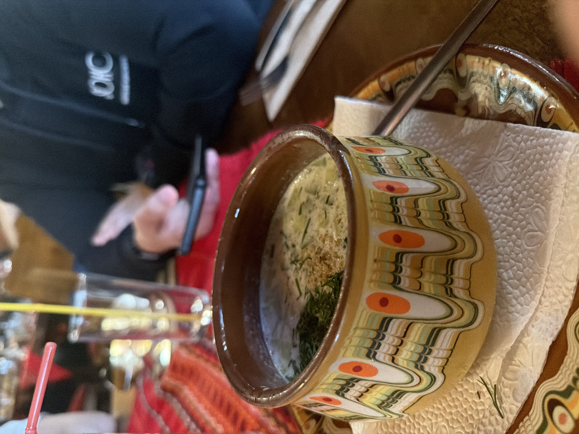 The image shows a bowl of Tarator, a cold soup popular in Bulgaria, placed on a plate with a decorative pattern, likely in a restaurant. The bowl is made of ceramic, with a tan and brown design, and contains a creamy white soup with green herbs, likely dill. A spoon rests in the bowl. The soup is served on top of a paper napkin on a plate, which is also decorated with similar patterns. In the background, there is a person wearing a black jacket with a logo, holding a mobile phone. There is also a glass with yellow and red straws, cutlery, and a table with a red and orange tablecloth. Given the presence of Tarator and the decorative patterns on the bowl and plate, the image is likely taken in Panagyurishte, Bulgaria, as this style of pottery is characteristic of the region. The scene suggests a dining experience in a traditional Bulgarian restaurant.