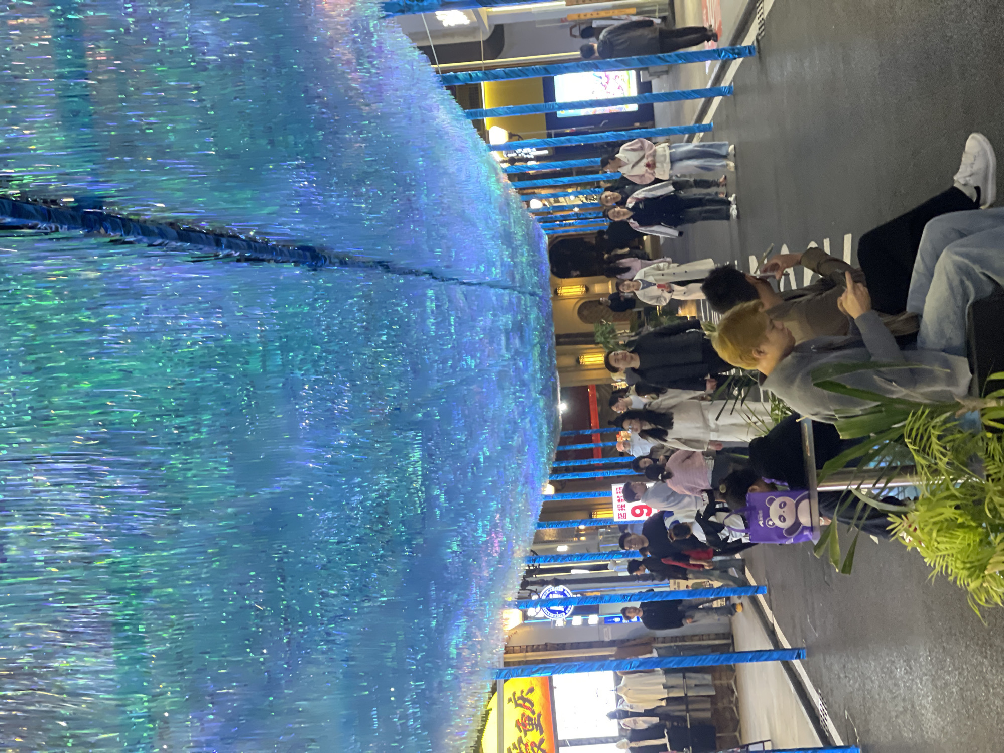 The scene depicts a pedestrianized street located within the Chongqing High-tech Zone, China, at night. An extensive overhead canopy, composed of dense, shimmering blue and green iridescent strips, spans the entire width of the street, providing the primary illumination. This canopy is supported by multiple vertical poles wrapped in blue material, delineating the pedestrian path. The ground is a dark paved surface, possibly asphalt, with white lines marking its edges. Storefronts line both sides of the street, displaying merchandise and emitting ambient light. A yellow sign on the left side prominently features the Chinese characters "爱重庆" (Love Chongqing).
Approximately 25 individuals are visible. The majority are pedestrians walking in various directions along the street, dressed in casual attire. In the immediate foreground, two males are seated on a bench or ledge to the right, oriented towards the right side of the frame. One male, with distinct light-colored hair, is actively engaged with a mobile phone. The male seated next to him is also holding a phone to his ear or mouth, possibly in conversation or using an earpiece. A purple bag adorned with a panda graphic is situated near the seated individuals. On the left, a storefront displays clothing items, including white shirts and jackets, on racks or mannequins. The environment is bustling with activity under the distinctive blue-green overhead lighting.