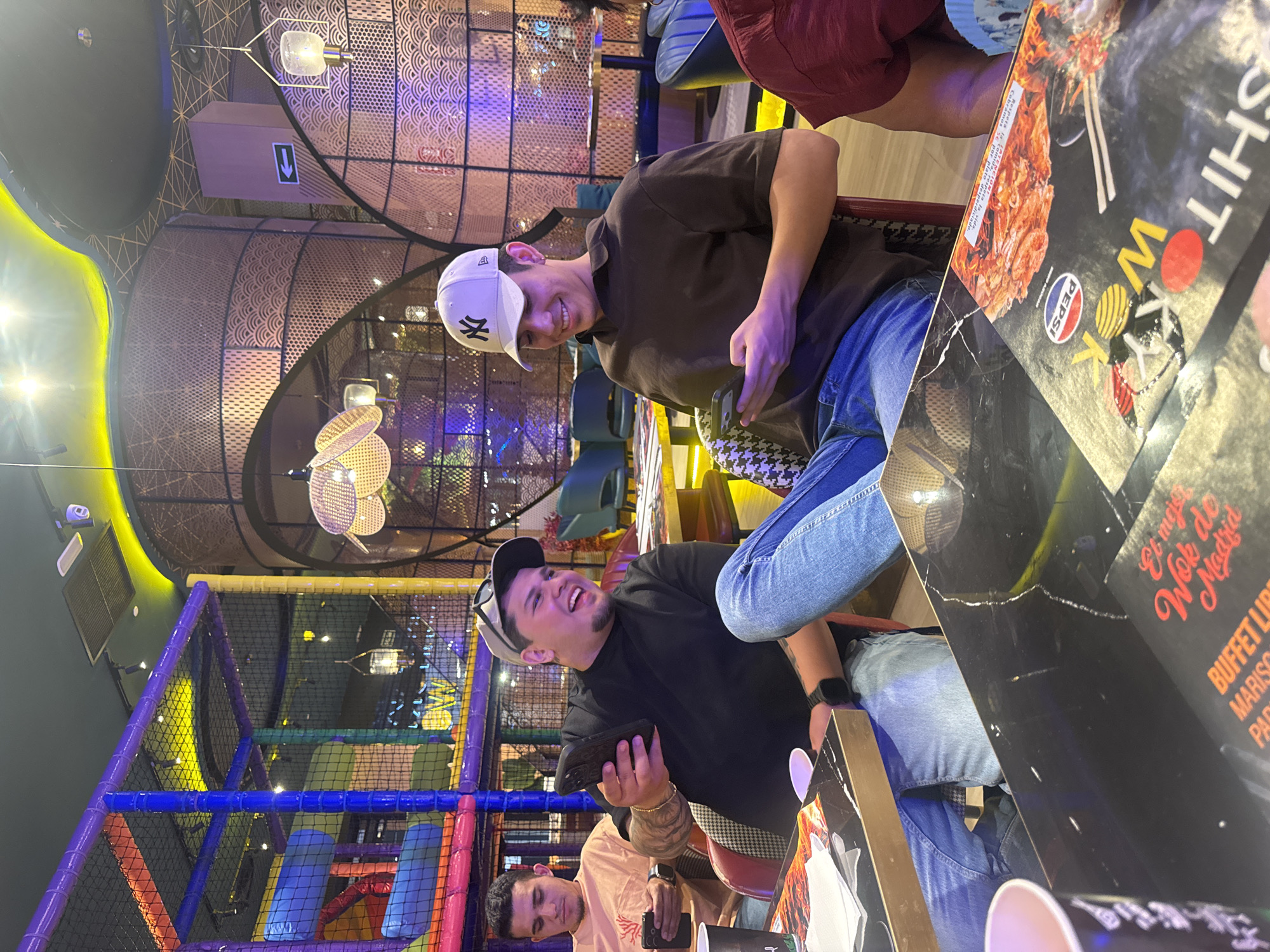 This is a vertical, medium shot photograph taken inside a restaurant. The primary subjects are three young men, two of whom are clearly visible, and a portion of a third. The man in the foreground, seated on the right, is wearing a black t-shirt and a baseball cap, and is laughing while holding a smartphone. He has tattoos on his arm. To his left, another man in a brown t-shirt and a white baseball cap is also smiling and holding a smartphone. Behind them, a third man in a light-colored t-shirt can be seen looking down at his phone. The setting appears to be a modern restaurant with elaborate decor, featuring metallic screens with intricate patterns and colorful ambient lighting. In the background, a brightly lit children's play area with blue, purple, and yellow netting is visible. A menu or placemat with text in Spanish, including "Et mej Wo Madria" and "BUFFET LIB," is spread across a table in the foreground. The overall mood seems casual and jovial, suggesting a social gathering, possibly a family outing or a meal with friends. The lighting indicates it is likely evening. The text on the menu suggests the restaurant is located in Madrid, Spain.