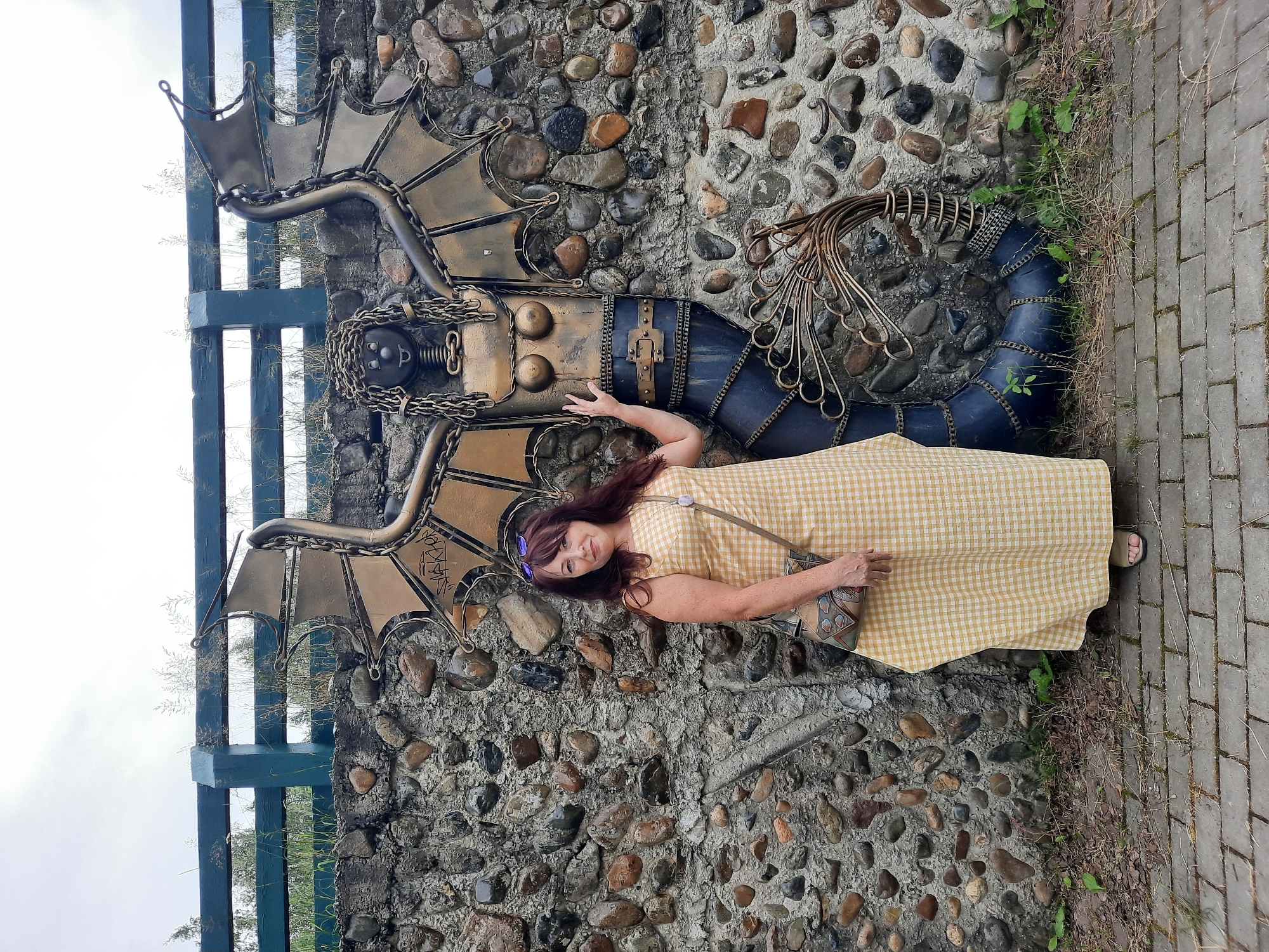 A woman stands next to a metal sculpture of a winged mermaid in front of a stone wall in Zelengorsky, Russia. The woman is wearing a yellow checkered dress and carrying a patterned bag. The sculpture is dark blue and gold, with a segmented tail and metal wings. The wall is made of rough stones and cement, and a blue wooden fence is visible at the top of the image. The ground is paved with bricks, and there is some dry grass and small plants around the base of the wall. The sky is overcast.