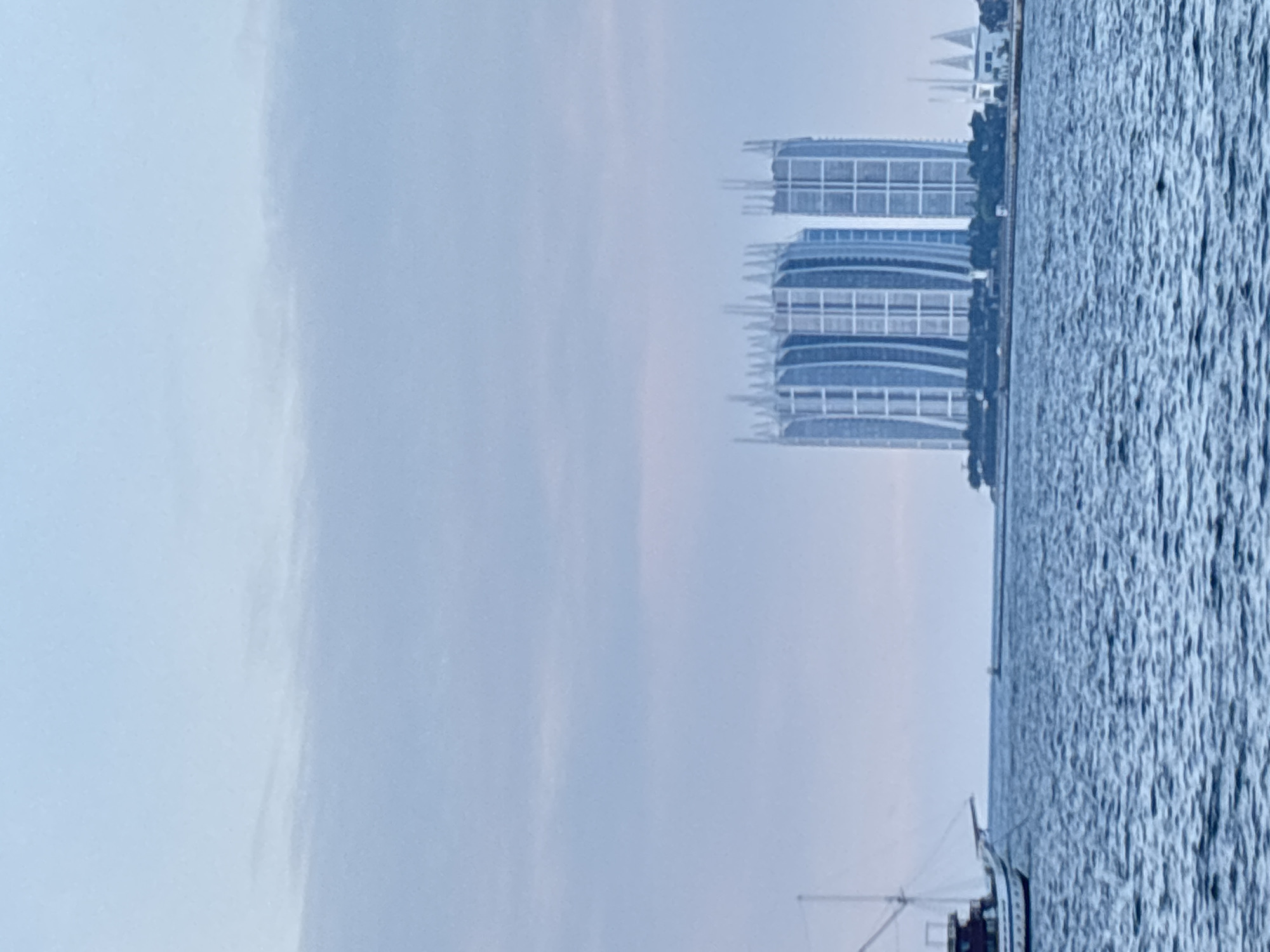 This image shows a body of water in front of several buildings in North Jakarta, Indonesia. The water is choppy and appears to be an ocean or large bay. In the foreground, a white boat with a dark hull and a mast is visible on the left side of the image. In the background, two large, modern buildings with curved facades and a distinctive lattice-like structure on top dominate the skyline. To the right of these buildings, a white structure with triangular peaks, possibly a building or part of a larger complex, is also visible. The sky is overcast and pale blue, with some faint clouds. The overall scene suggests an urban coastal environment.