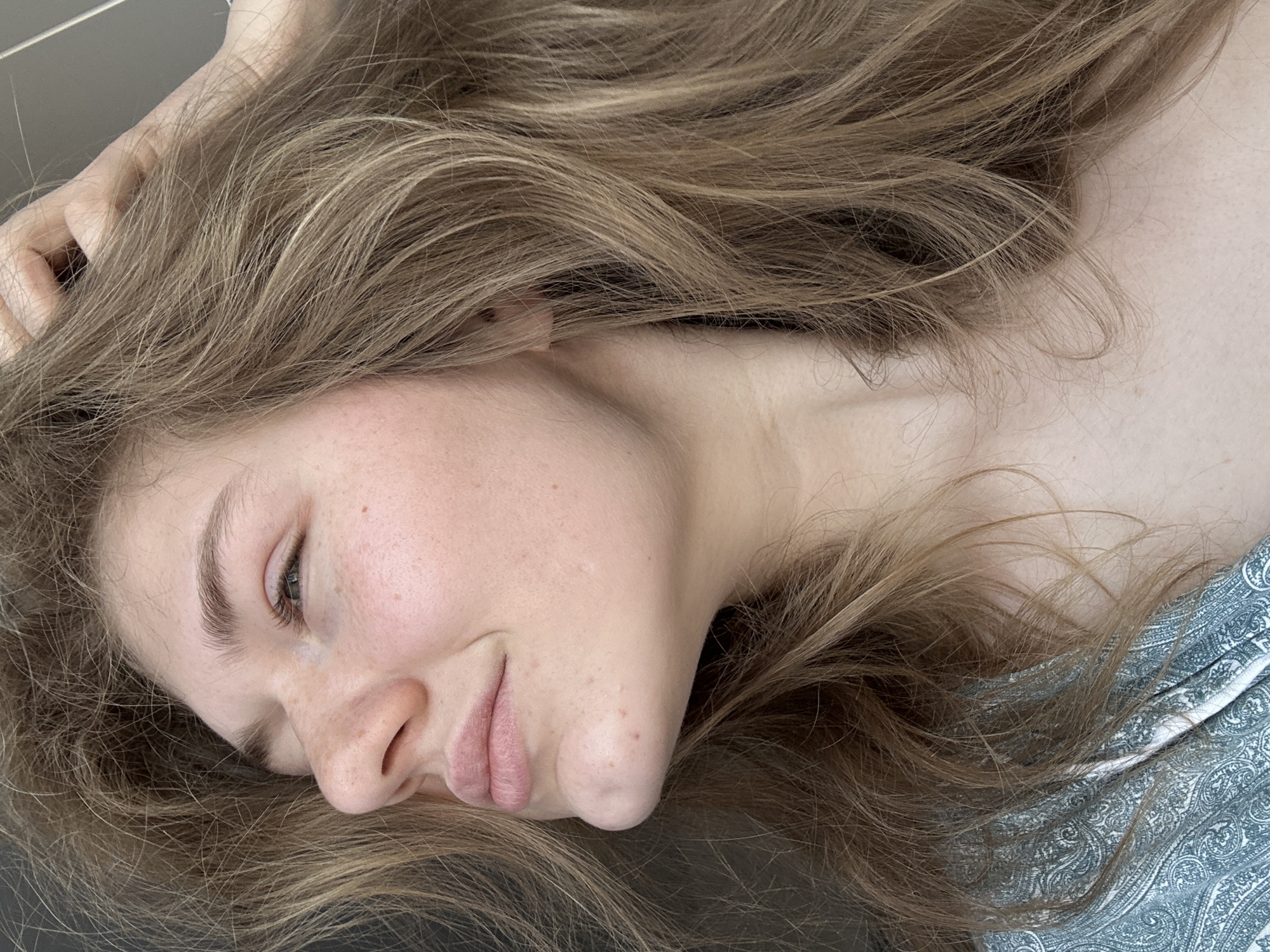 A young woman is lying down, facing slightly to the left. She has light skin, freckles, and light brown hair that is spread around her head and neck. Her eyes are partially closed, and her lips are slightly parted. She is wearing a paisley-patterned top. The background is a flat, dark surface. There is no indication of location (city of Kazan, Russia, or otherwise).