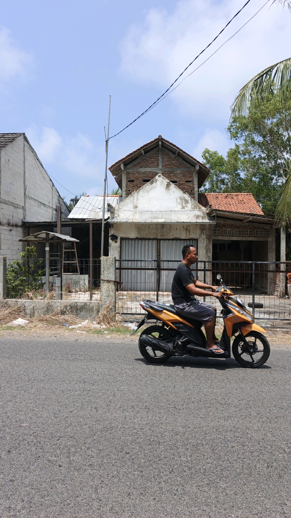 A man rides an orange scooter down a paved road in Desa Karangjaladri, Indonesia. The man wears a black t-shirt and grey shorts, with his bare feet in flip-flops. He is riding past a partially constructed building with a red tile roof and bare brick walls. The building has a large corrugated metal garage door. In front of the building is a metal fence with a gate. To the left of the building is another unfinished structure with white walls. Palm trees and other foliage are visible behind the buildings. The sky is blue with scattered clouds.