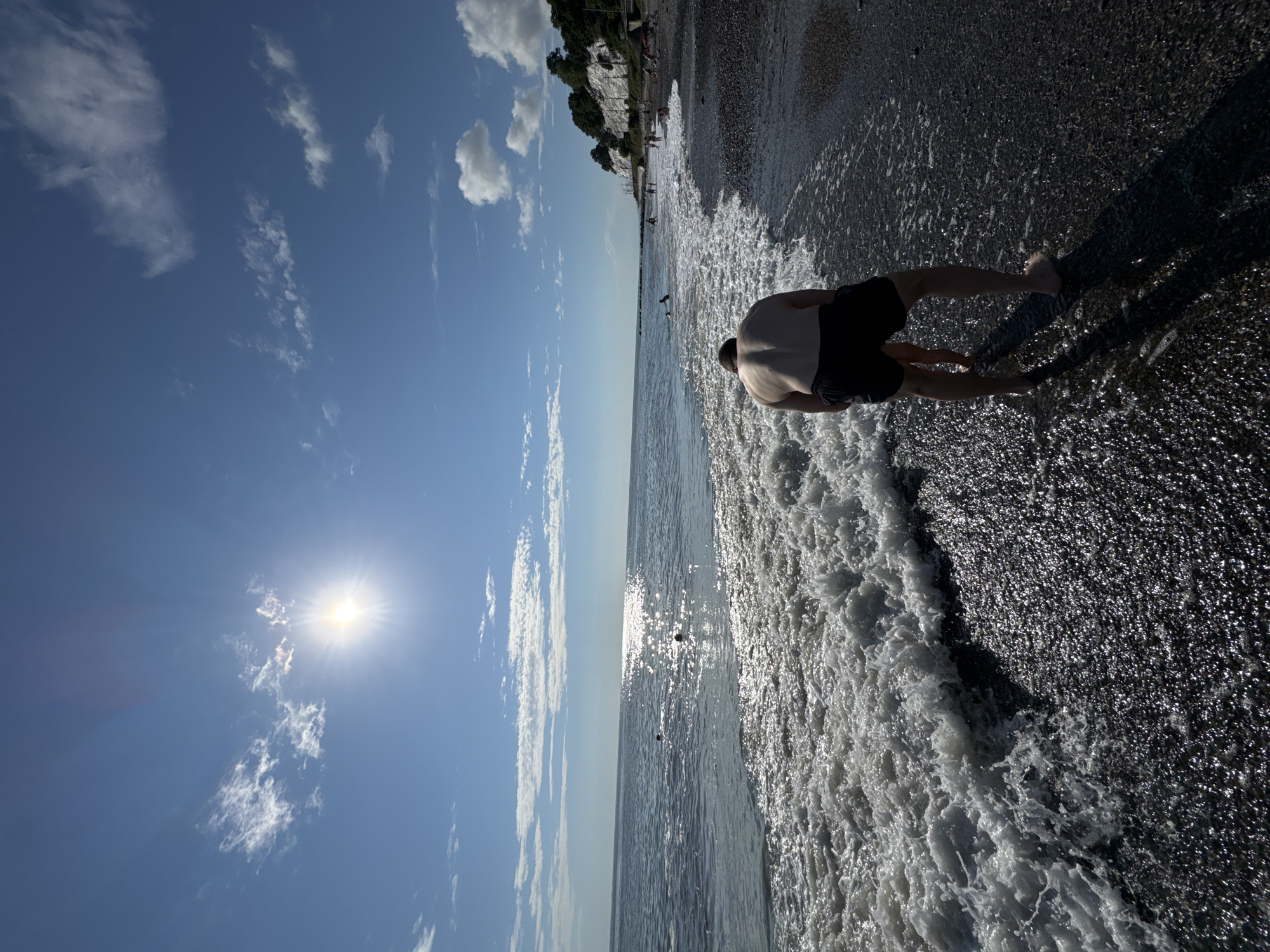 An overhead shot captures a person in black shorts facing away from the camera, standing on a pebble beach at the edge of the Black Sea. The water, with a frothy edge, washes over the pebbles. In the distance, other people are visible swimming and sunbathing along the shoreline, which is backed by a cliffside covered in greenery. The sky above is a clear blue, dotted with scattered white clouds, and the sun shines brightly. The scene appears to be a typical summer day at a beach resort in Sochi, Russia, with the sun's glare reflecting off the water.