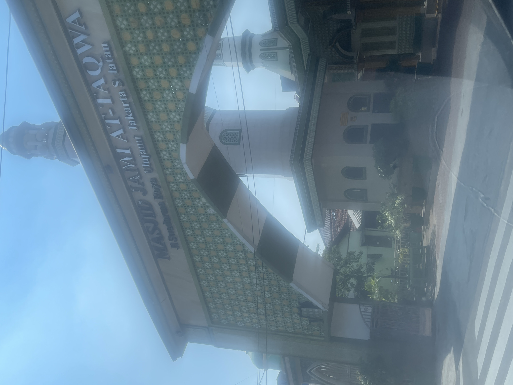 The image shows the entrance of Masjid Jami At-Taqwa, located on Jalan Swadarma Raya in South Jakarta, Indonesia. The mosque has a grand archway with intricate green and gold patterned screens. Above the archway, the name of the mosque is inscribed in bold, silver lettering: "MASJID JAMI AT-TAQWA". Below this, smaller text reads "Jl. Swadarma Raya Ulujami Jakarta S Jatan". The mosque's minarets are visible in the background, with one taller and more prominent than the other. The sky is clear and blue, suggesting a sunny day. In the foreground, a portion of a street with white pedestrian crossing lines is visible, indicating the mosque is situated by a road. Lush green trees and some buildings are visible behind the mosque entrance. The overall scene depicts a prominent religious building in a city environment.