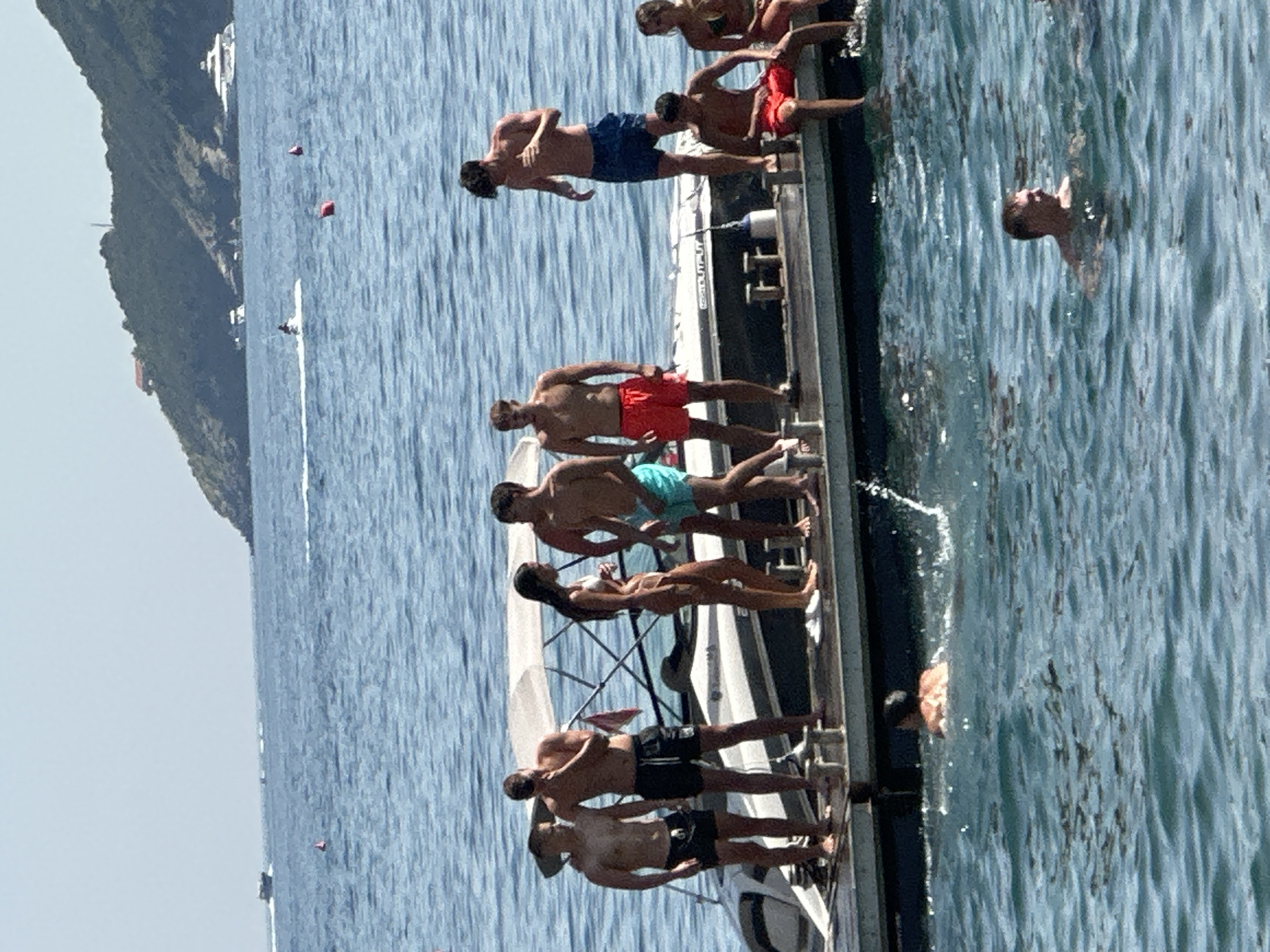 A group of eight young adults, dressed in swimwear, are standing and sitting on the deck of a boat or pier in the sea. The water is blue and appears calm. In the background, a green, hilly island with some buildings is visible. Several boats, including a yacht and a jet ski, are also present in the distance. The sky is clear and blue. The scene suggests a sunny day in a coastal recreational area, likely Budva, Montenegro, given the presence of a jetty and the characteristic landscape.