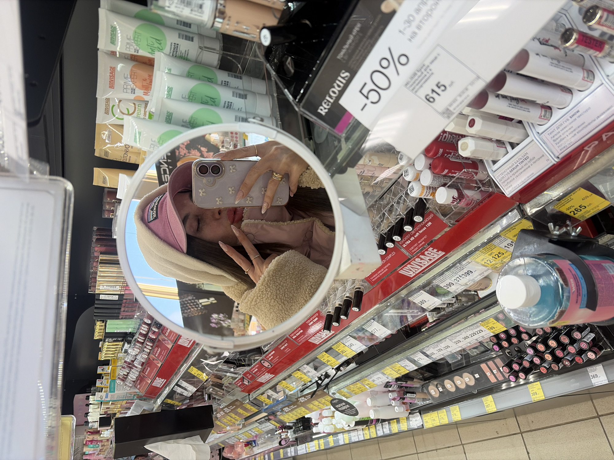 The image shows a selfie of a young woman reflected in a circular mirror in a store. She is wearing a pink hat and a beige jacket. She is holding a smartphone with a lavender-colored case decorated with small flowers. Her nails are long and decorated. The mirror is reflecting a shelf in a store, filled with various cosmetics products, including lipsticks, concealers, and lotions. A sign above the products indicates a 50% discount. The text on the products is in Russian, and the prices are listed in Russian Rubles. Based on the brands and the overall appearance of the store, it is likely located in Saint Petersburg, Russia.