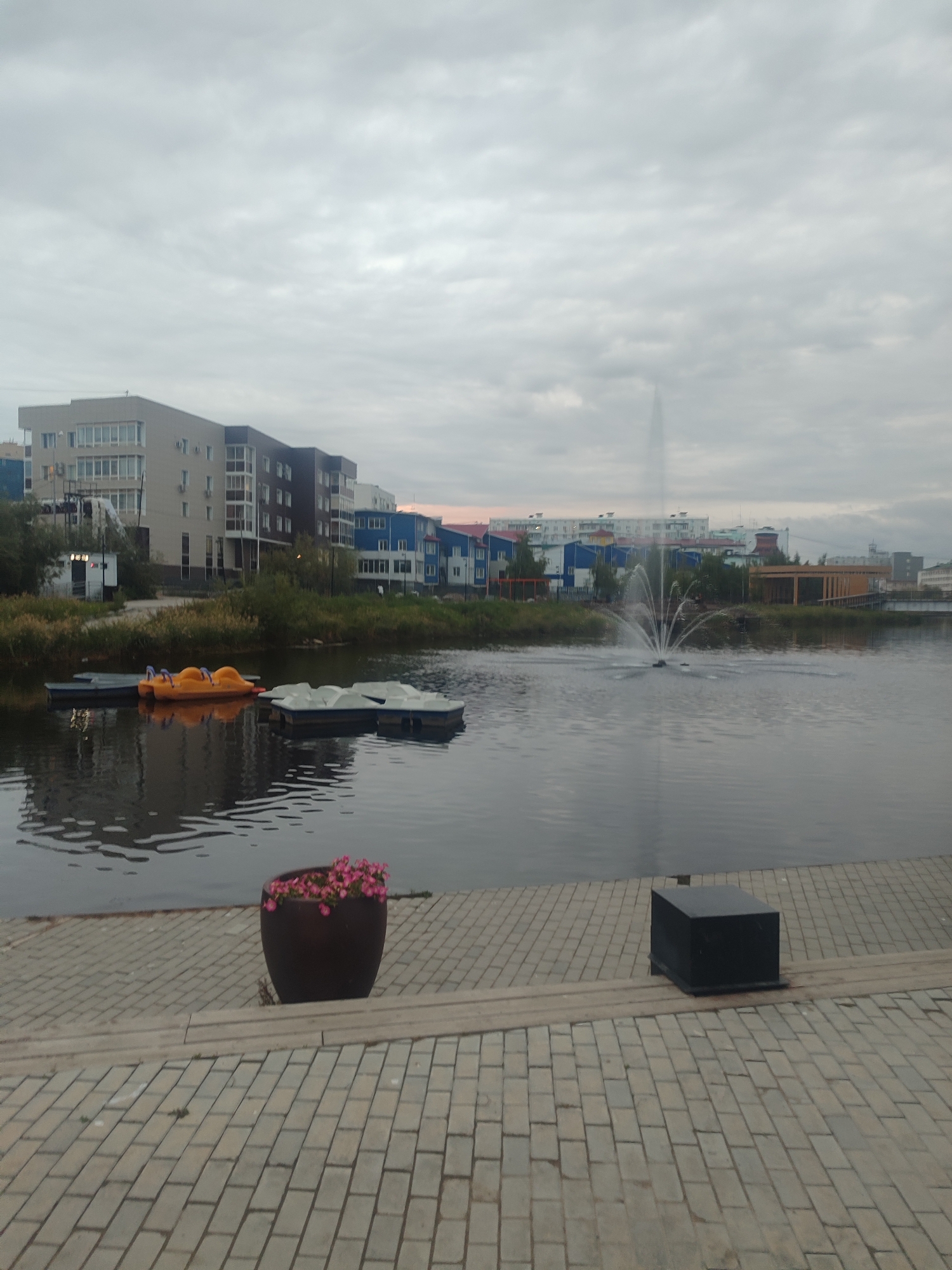 The image depicts a cloudy day at a waterfront in Yakutsk, Russia. In the foreground, a paved walkway leads to the water's edge, where a large, dark planter filled with pink flowers is visible. To the right of the planter, a black, cubic object rests on the pavement. The water features a fountain in the middle distance, with several pedal boats docked on the left side. Across the water, a line of modern apartment buildings, varying in color and height, forms the cityscape. The sky is overcast, with a dull, grey light illuminating the scene.