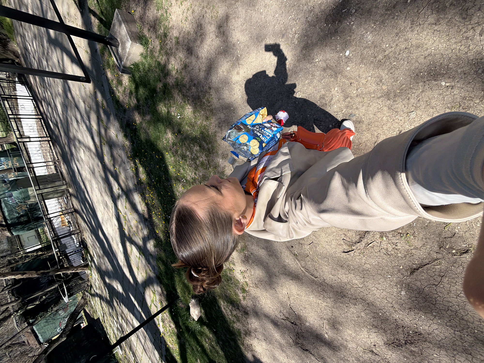 The image depicts a person standing on a dirt path in what appears to be a park. The person is holding a bag of chips and a small red and white pom-pom-like object. The person is wearing a light-colored jacket, a scarf, orange pants and white sneakers. Their shadow is visible on the path. The path is cracked and dry, with some grass and small yellow flowers on the edges. In the background, there is a metal fence and some trees. The picture was taken in Sofia, Bulgaria, in the park near the zoo.