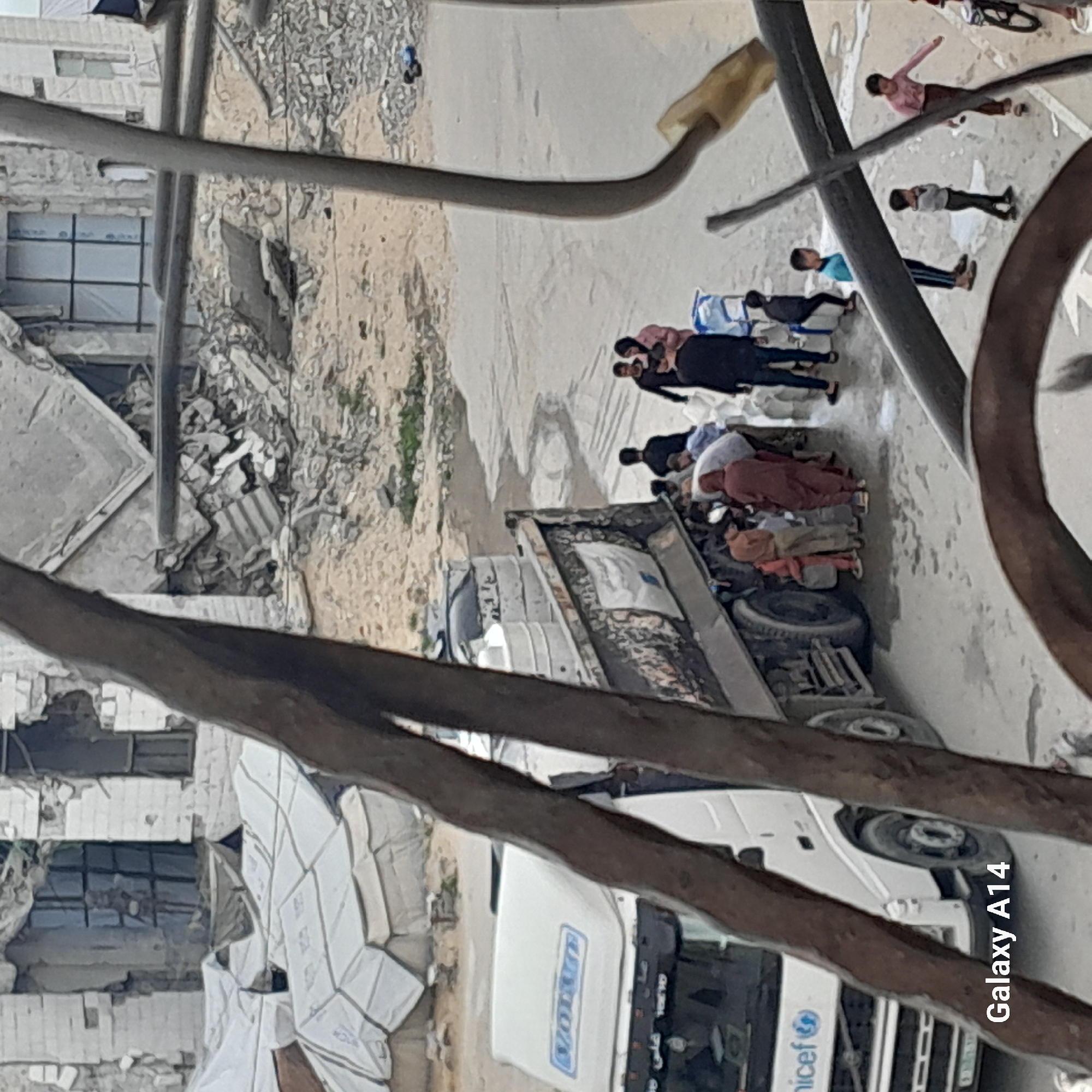 The image, taken from an elevated perspective and oriented vertically, captures an outdoor scene dominated by signs of destruction and humanitarian activity.
In the mid-ground, a large white truck with a flatbed is positioned on what appears to be a dirt or unpaved road. The truck clearly displays the "Unicef" logo on its side and the "VOLVO" brand on its cab. The truck's bed is filled with various items, possibly supplies. Around the truck, several adults are gathered; some women wear head coverings. They appear to be engaged in the distribution or collection of goods, with one person leaning into the truck bed and others standing nearby. White bags or sacks are visible near the truck, and some white barrels or containers are on the ground beside it.
Further away from the truck, several children are present, standing or walking, observing the activity or moving through the area.
To the left side of the image, the background reveals extensive damage. Piles of rubble, shattered concrete, and exposed rebar indicate severely damaged or destroyed buildings. Parts of walls with empty window frames are still standing amidst the debris, underscoring the devastation.
The foreground is occupied by dark, out-of-focus structural elements, possibly railings or wires, suggesting the photo was taken from a building or overhead structure. The time of day appears to be daytime with clear weather. The overall scene suggests a context of post-disaster or conflict relief efforts. The watermark "Galaxy A14" is visible in the bottom right corner of the original image, indicating the device used. Faint Arabic script is also visible on the truck's grille, with one word possibly reading "علي" (Ali).