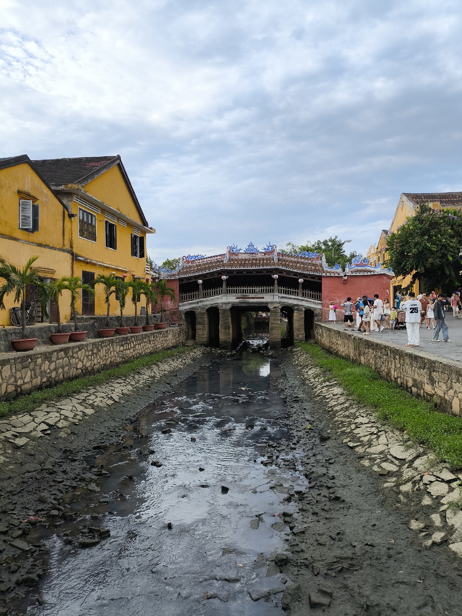 This image depicts a scene in Hoi An, Vietnam, featuring the iconic Japanese Covered Bridge. The foreground shows a low-water canal with exposed mudflats and scattered rocks, reflecting the overcast sky. The canal is lined with stone walls and patches of green grass.
To the left, a yellow building with a dark tiled roof and visible windows stands prominently. In front of this building, a row of potted palm trees is arranged along the stone wall.
The Japanese Covered Bridge, a historically significant landmark, is centrally located, spanning the canal. It has a traditional tiled roof with decorative elements. The bridge is supported by stone pillars and has a walkway with railings.
On the right side of the canal, a group of people are walking along the pathway beside the bridge. They are dressed in casual clothing, and some are carrying bags. One individual is wearing a white outfit with the number "25" visible. The pathway is bordered by a stone wall and green vegetation. Further to the right, another yellow building with a red section is visible, with trees providing shade. The sky is cloudy, indicating an overcast day. The overall scene suggests a tourist area within the city.