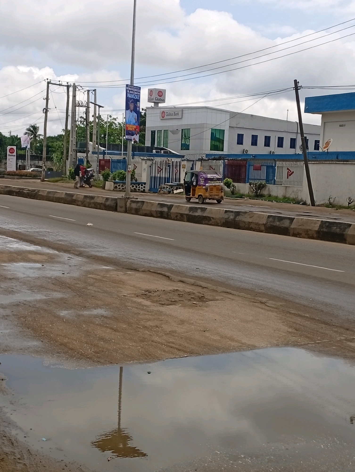 This is a medium-shot, eye-level, outdoor photograph taken on a cloudy day, likely in Ikeja, Nigeria. The scene captures a street with a Globus Bank building in the background, identified by signage. A tall lamp post stands prominently in the foreground, with its reflection visible in a large puddle on the road. A yellow and purple auto-rickshaw, also known as a Keke Napep, is visible driving on the road. A man on a motorcycle is stopped near the sidewalk, and another person is walking on the sidewalk. A political poster featuring Dr. Olajide Hamzat for Governor in 2027 is attached to the lamp post. The road appears to be a bit wet, suggesting recent rain. The overall environment suggests a typical urban setting in Nigeria.