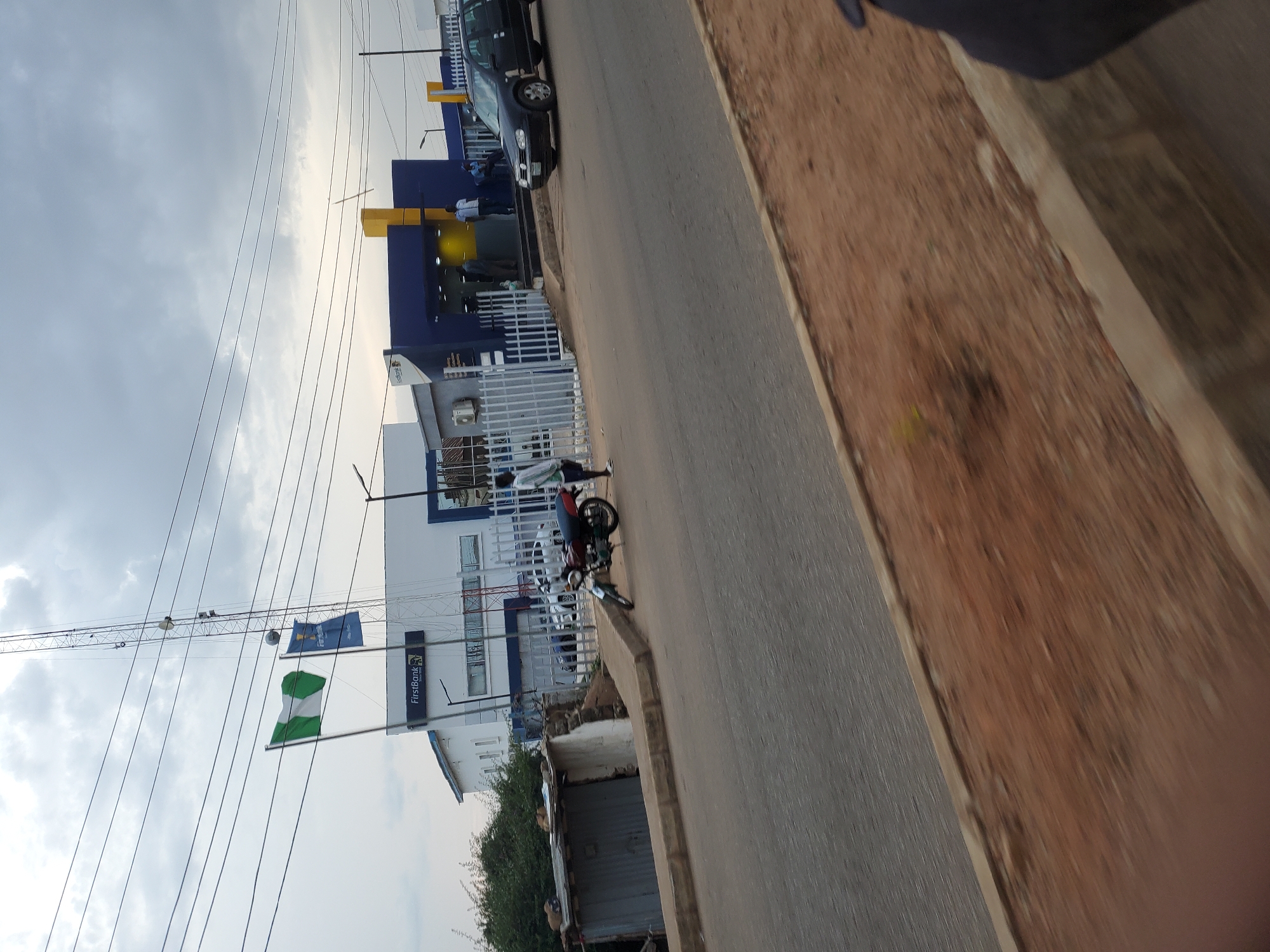 This is a photograph taken at street level in Bukuru, Nigeria. The scene depicts a roadside view with a building prominently featuring the "FirstBank" logo. A Nigerian flag is visible, suggesting it's a public building or an official event is taking place. The building itself is modern, with blue and white accents and a white metal fence.
In front of the building, several people are gathered, some standing near a dark-colored car. A motorcycle is parked on the side of the road. The street is paved, and to the right of the road, there is a brown, somewhat rough, embankment. The sky is cloudy, indicating it might be late afternoon or early morning, as the light is diffused. Power lines crisscross the sky, a common sight in urban areas of Nigeria.
A taller structure, possibly a crane or a telecommunications tower, is visible in the background, extending above the building. The overall atmosphere appears to be one of everyday activity in a commercial area. There's no strong emotional tone conveyed, but the presence of the bank and people suggests normal business operations. The ground beside the road is covered with reddish-brown earth, typical of the soil in many parts of Nigeria.