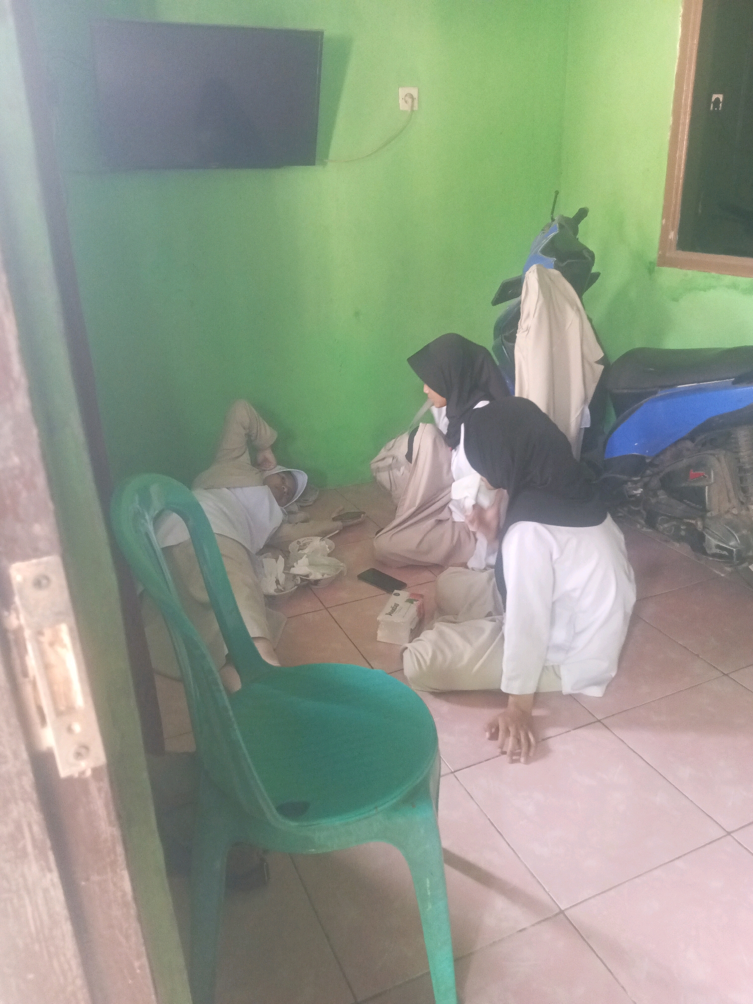 In Purwakarta, Indonesia, the image captures a scene within what appears to be a simple room with light green walls. A flat-screen television is mounted on the wall. On the floor, three individuals are present, all dressed in light-colored uniforms with headscarves. Two of them are seated or kneeling, with one wearing a black headscarf and the other a light-colored one. The third individual is lying down, apparently asleep, with their head resting on a white fabric. A green plastic chair is positioned in the foreground, partially obscuring the lower portion of the scene. A blue motorcycle is visible in the background, with a beige garment draped over its handlebars. The floor is tiled in a pale pinkish-brown. The overall impression is of a shared living or resting space, possibly in a school or dormitory context, given the uniforms.