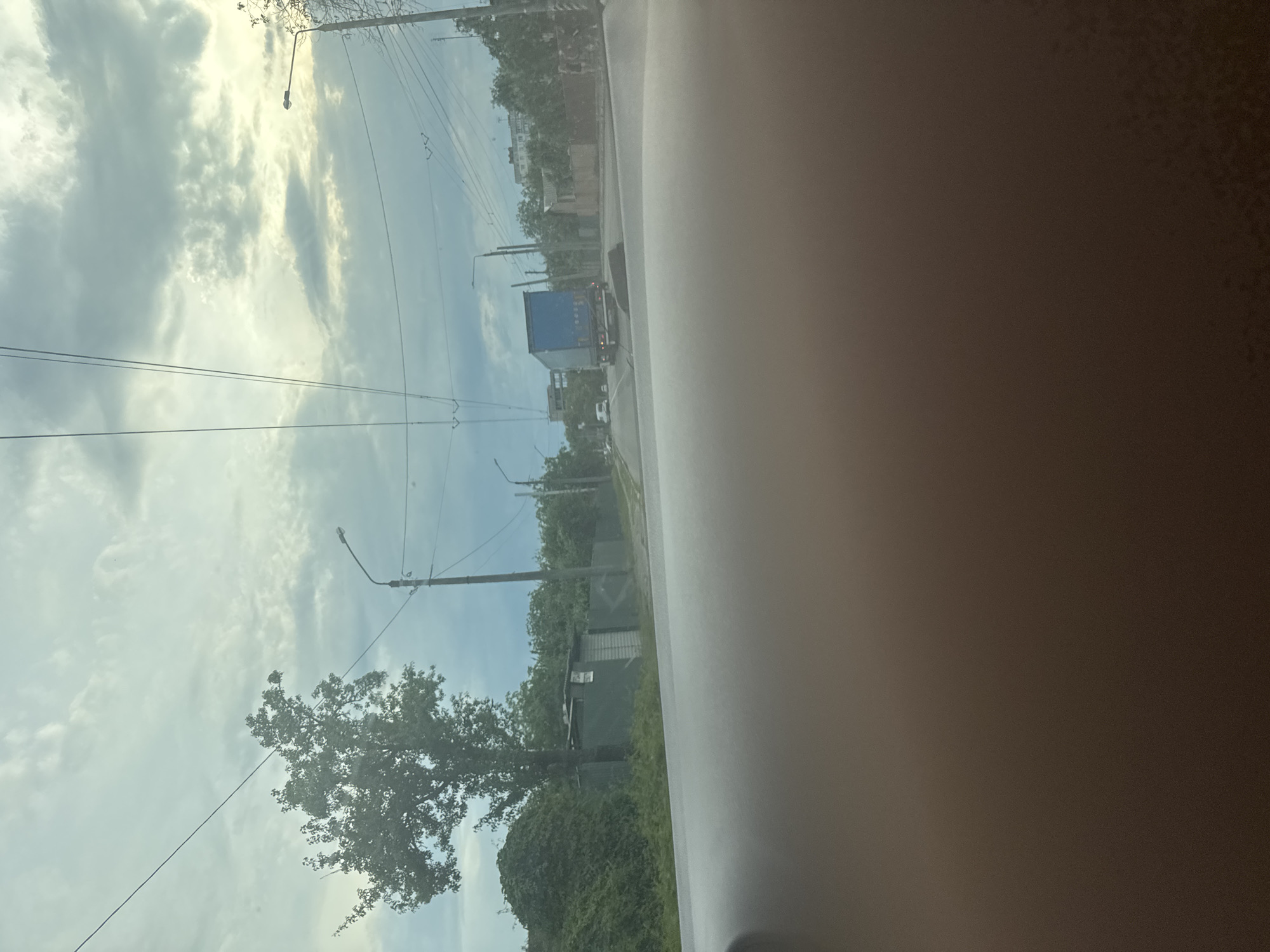The scene depicts an outdoor street or road environment, viewed from the interior of a vehicle, in the city of Unknown, Unknown. The lower approximately 40% of the image is occupied by an out-of-focus, dark brown, textured surface, likely a vehicle's dashboard. The sky above is heavily overcast with a mix of light gray and white clouds.
A paved asphalt road, appearing dry, extends into the distance. On the left side of the road, a large deciduous tree with dense green foliage is prominent. Behind the tree, a mass of green vegetation, possibly vines, covers a sloped area. Further along the left, a long green corrugated metal fence runs parallel to the road, featuring a small, box-like structure also constructed from corrugated metal, with a white rectangular sign affixed to it.
Multiple utility poles stand along both sides of the road, supporting a complex network of overhead wires, including what appear to be power or trolleybus lines. Streetlights are mounted on several of these poles. A blue semi-trailer truck is observed on the right side of the road, oriented away from the viewer, with its rear lights visible. Further down the road, at least two smaller white passenger vehicles are discernible. In the far background, beyond the trees and utility lines, the faint outlines of multi-story buildings are visible. No individuals are distinctly identifiable in the frame. The lighting suggests daytime under an overcast sky.