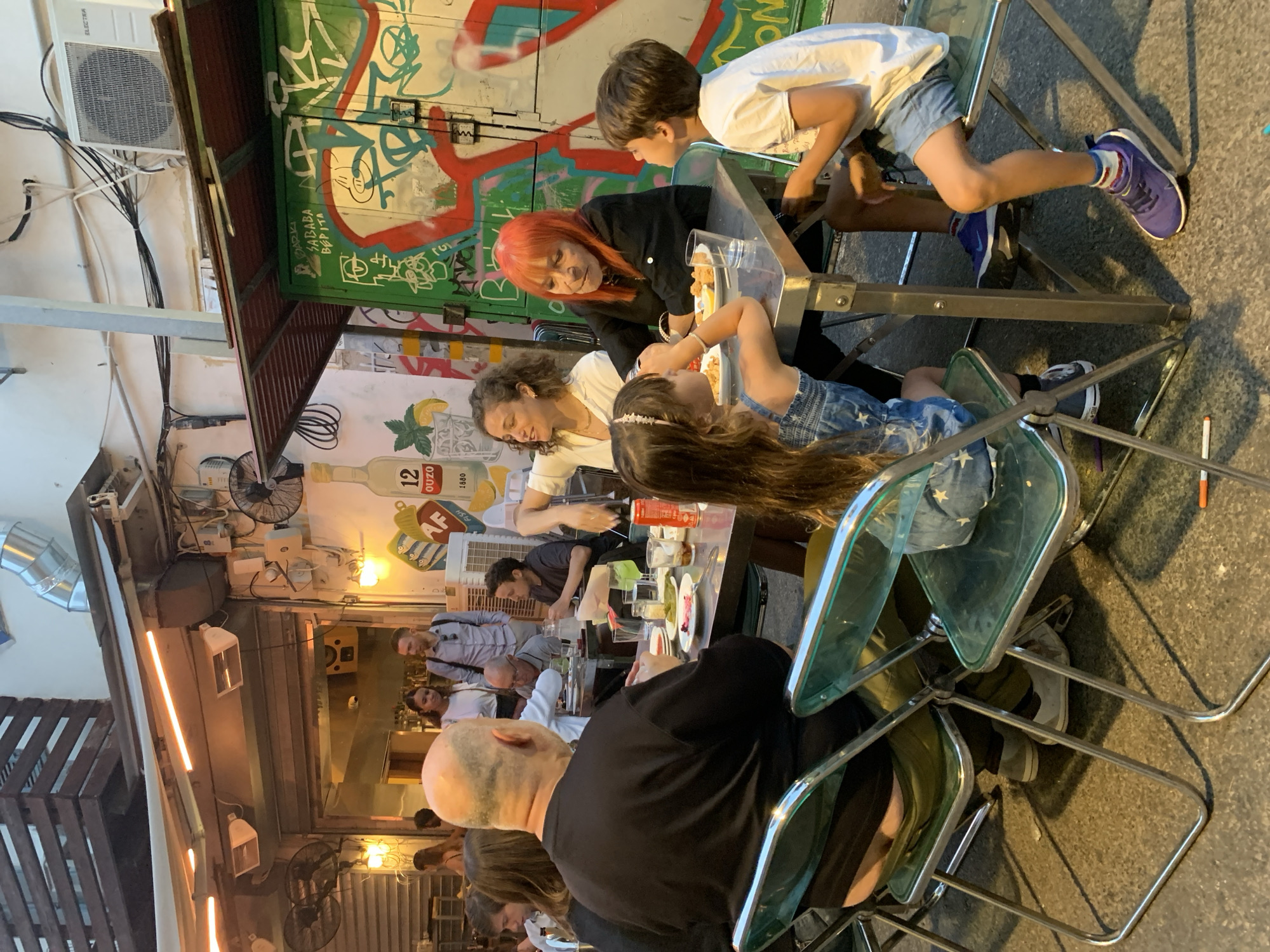 The image captures a social gathering in an outdoor dining area, likely in Tel Aviv, Israel, given the presence of the "12 Ouzo" brand bottle, commonly found in Greek and Israeli establishments. A group of people are seated at tables, enjoying a meal or conversation. A woman with red hair interacts with a young boy, while another woman engages with a young girl at an adjacent table. The tables are set with plates, glasses, and bottles. The surrounding environment is characterized by graffiti-covered walls, air conditioning units, exposed wiring, and industrial-style lighting, creating an urban, somewhat gritty atmosphere. The scene is well-lit, suggesting either late afternoon or early evening. The chairs are a light-green tinted plastic with a thin silver metal frame.