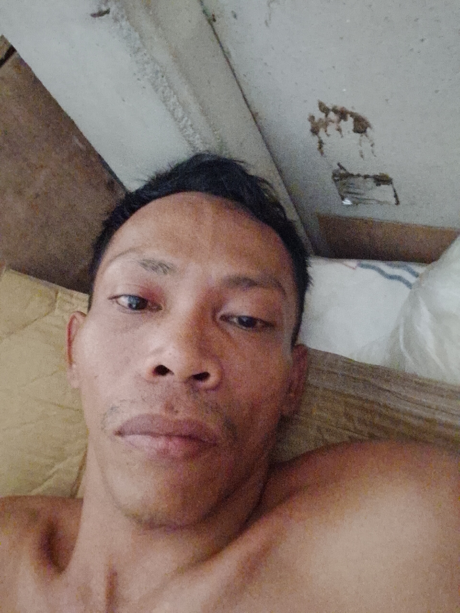 A close-up selfie taken indoors, likely in Bukit Ketok, Indonesia, shows a man lying down. His head rests on a tan pillow next to a white pillow. He is facing the camera with a neutral expression. His hair is short and black. The background includes a wooden surface and a white ceiling with stains.