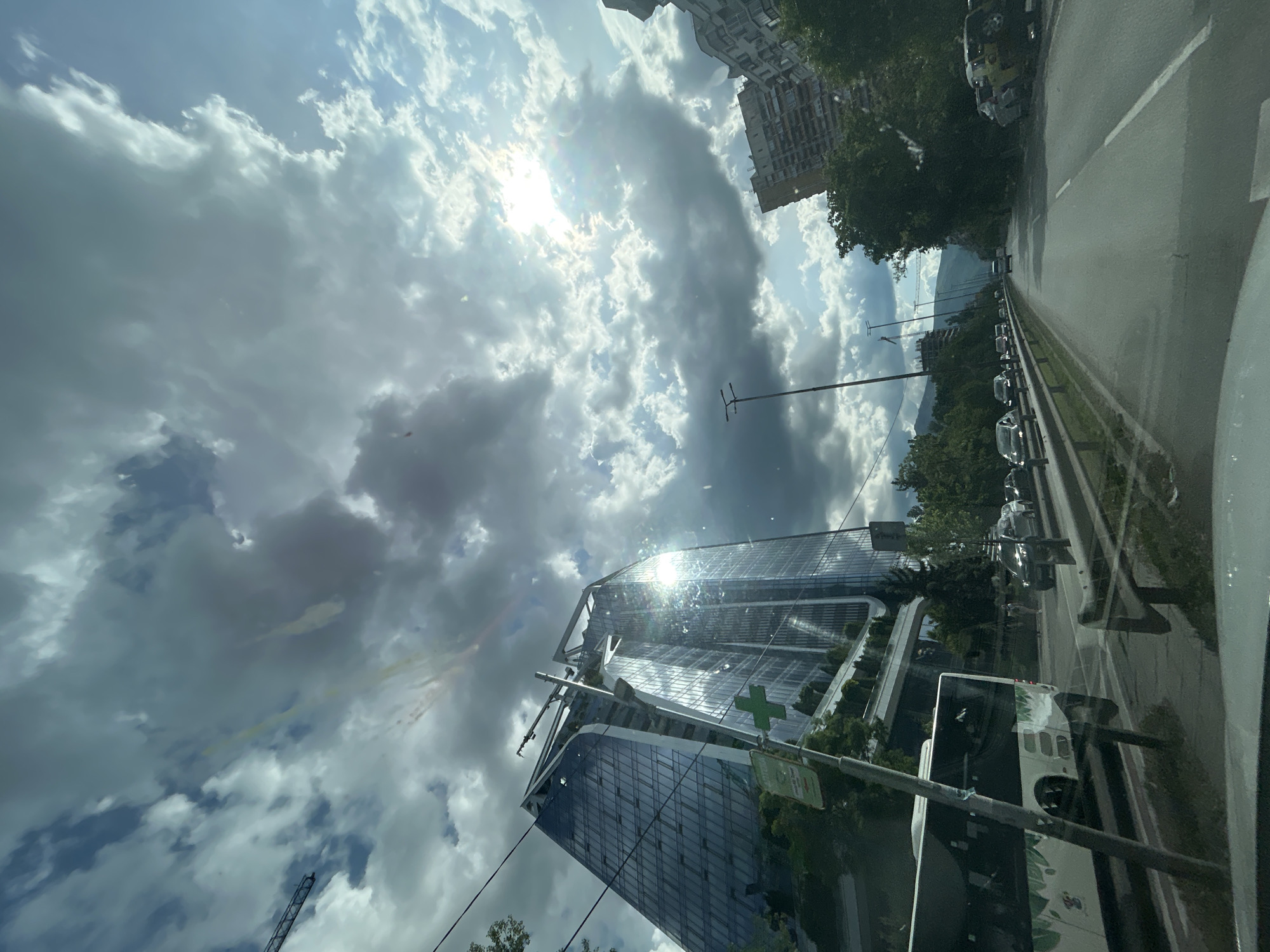 The image, taken from inside a vehicle, depicts a daytime scene in Sofia, Bulgaria. A modern glass building with a distinctive architectural design dominates the lower left portion of the frame. Sunlight is reflected off its glass facade. In the background, a cloudy sky with the sun obscured partially by clouds. A road with cars parked and moving alongside the building. A bus is located near the bottom of the building.