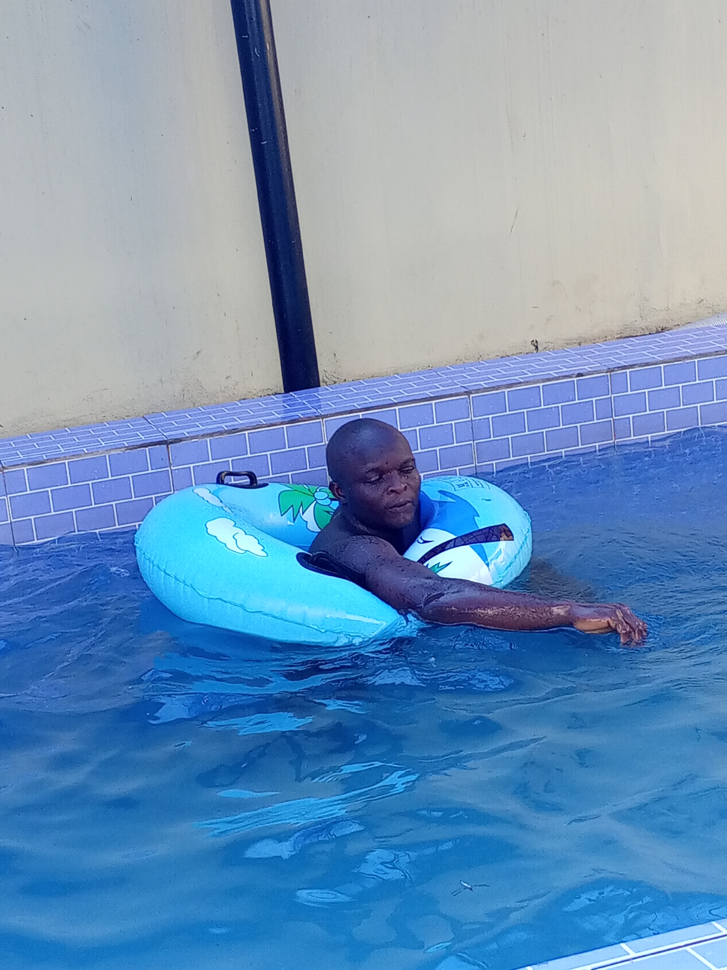 The image depicts a man floating in a swimming pool in Ankpa, Nigeria. The man is using a blue inflatable tube with handles and graphics of clouds and a palm tree. The pool is surrounded by blue and white tiles, and a beige wall is visible in the background, along with a black pole. The water is a clear blue, and the man has one arm extended into the water.