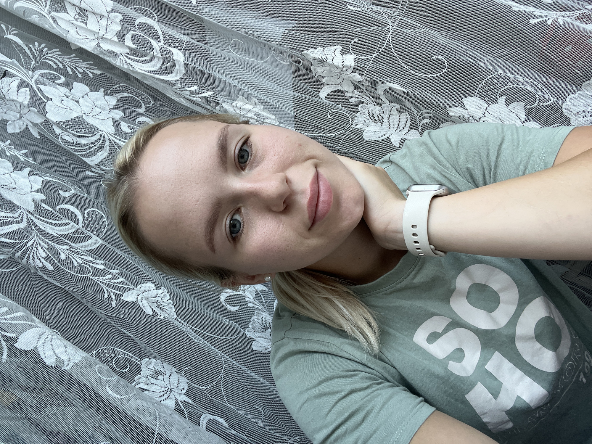 A young, blonde woman is shown in a close-up selfie. She is wearing a light green t-shirt with white lettering on it that appears to say "SO". She has her right hand resting on her chin and her left arm is visible with a white watch on her wrist. She has light blue eyes and a slight smile. Behind her, a white lace curtain with a floral pattern is draped. The scene appears to be indoors, with no discernible city or specific location information available.