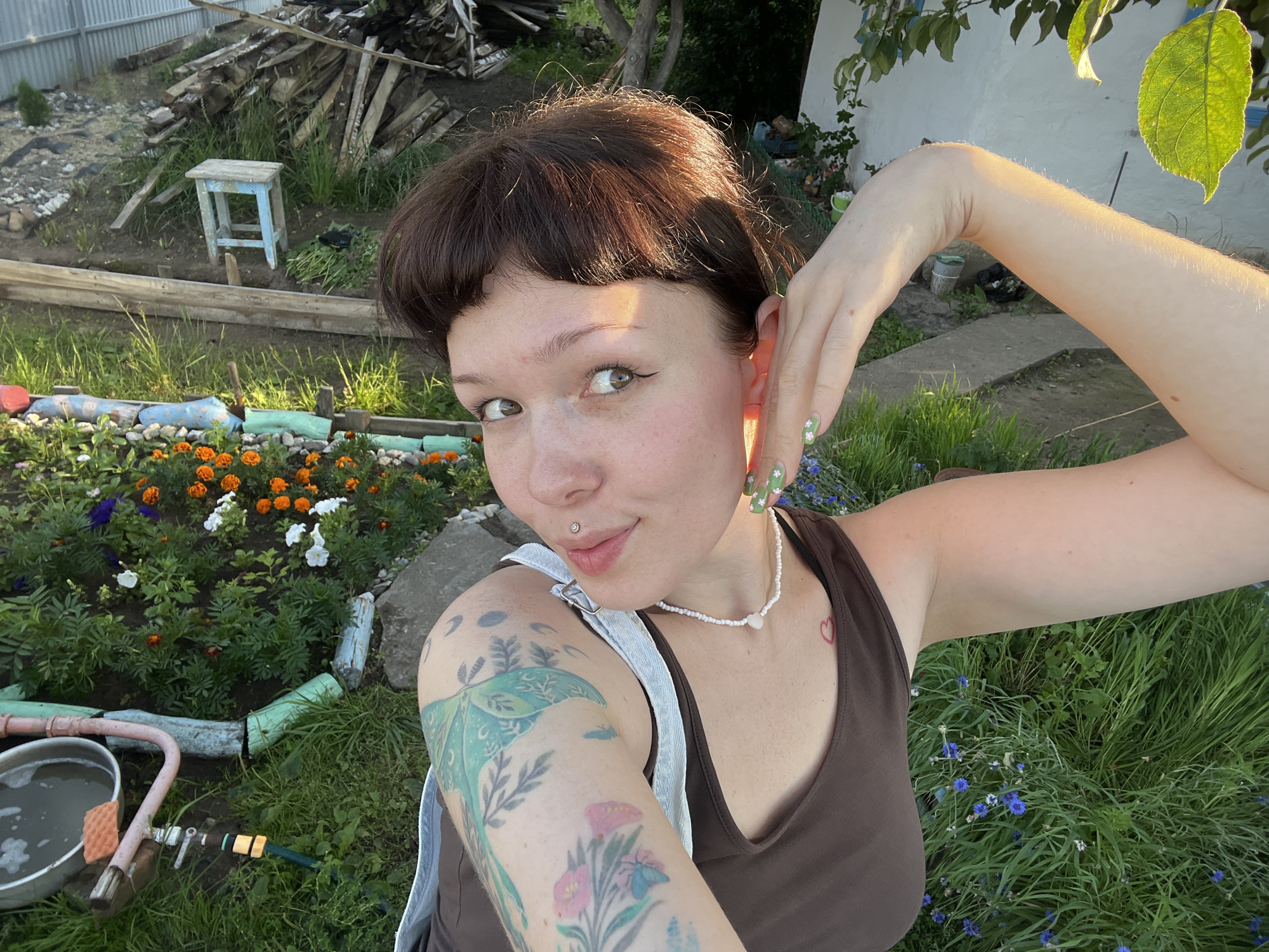 A young Caucasian female with dark brown hair and fair skin is taking a selfie outdoors in a garden. She is wearing a brown tank top and a beaded necklace. Her right arm is raised, with her hand near her ear. She has a lip piercing and tattoos on her left arm. The background shows a flower bed with orange and purple flowers, a bucket with water, a garden hose, and a pile of lumber. The setting appears to be a backyard or a dacha, possibly in Chaikovsky, Russia, given the rustic elements and common practice of gardening in such locations. The lighting suggests it is late afternoon or early evening.
