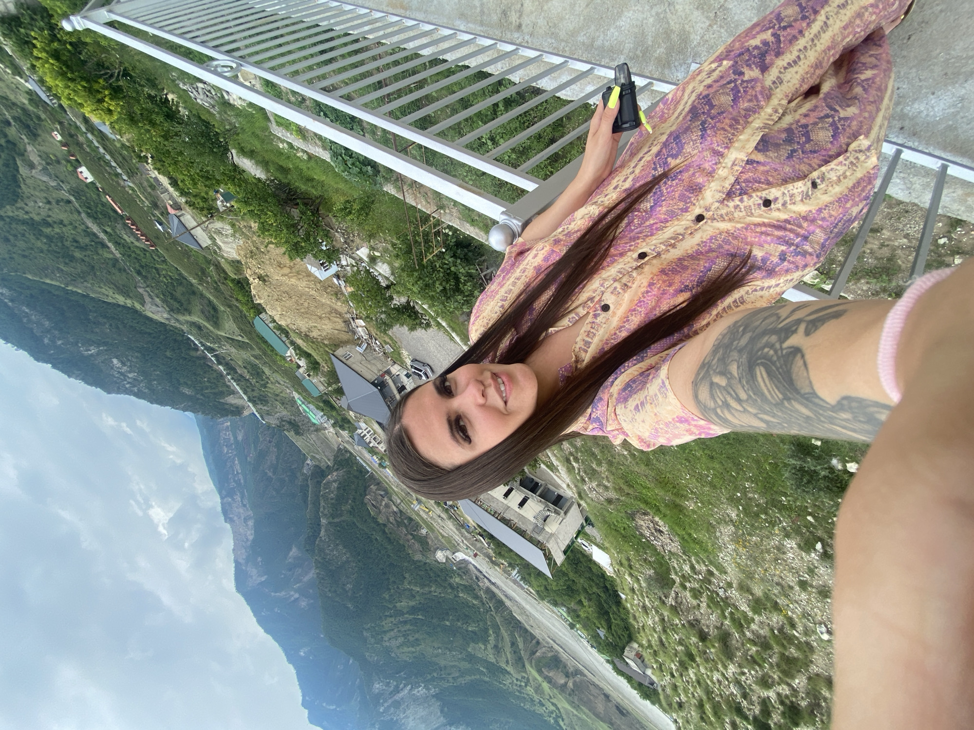 In Chalda, Russia, a woman with long dark hair and a tattoo on her arm takes a selfie outdoors. She is wearing a dress with a snake-like pattern in shades of pink and purple. In her right hand, she holds a black object with bright yellow fingernails. She stands on a balcony with a metal railing, overlooking a valley. The landscape features steep, green mountains, a river, and scattered buildings and houses along the slopes. The sky is overcast.