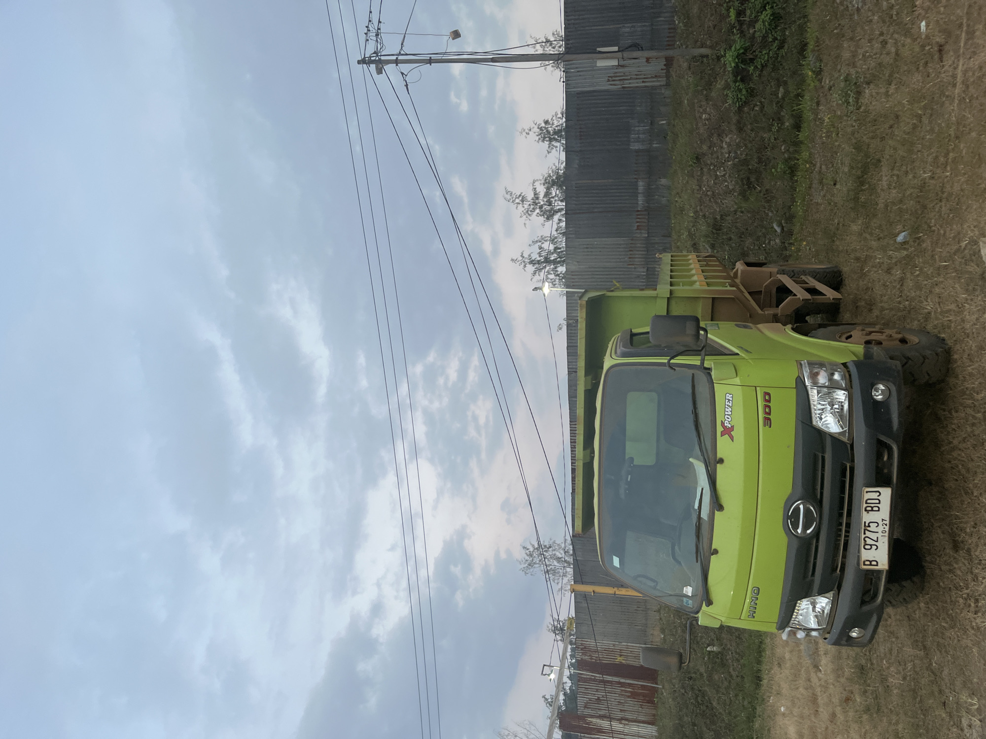An eye-level, medium shot captures a bright green Hino dump truck parked on dry, grassy terrain under an overcast sky. The truck is positioned facing forward and slightly to the right. Its front features a black grille with a silver Hino logo, clear headlights, and a license plate reading "B 9275 BDJ 10-27". The truck's body has "HINO", "XPOWER", and "300" branding. Behind the truck, a corrugated metal fence runs horizontally, with a utility pole and power lines stretching across the upper portion of the frame. Trees with sparse foliage are visible behind the fence, and the ground is covered in dry grass and scattered debris. The scene suggests an outdoor industrial or undeveloped area.