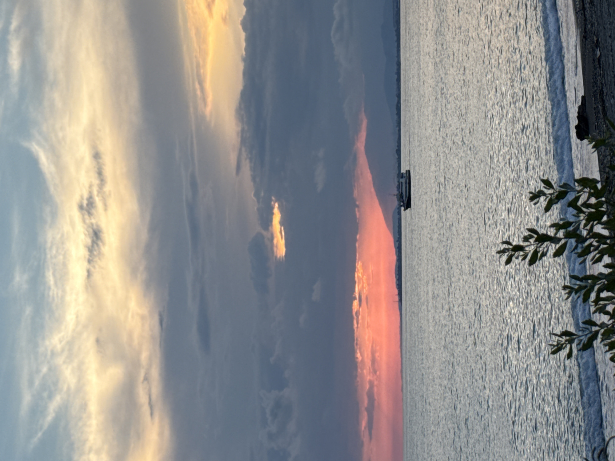 A serene sunset over the sea in Yuada-Gulrypsh, Abkhazia. The sky is a canvas of dramatic clouds, with vibrant hues of pink and orange from the setting sun peeking through darker grey clouds. A sliver of sunlight breaks through the clouds in the upper right, casting a warm glow. In the distance, a silhouette of mountains forms the horizon. A small boat sails calmly on the gently rippling water. In the foreground, the edge of a beach is visible with small waves lapping the shore, partially obscured by the branches of a leafy plant. The overall impression is one of tranquility and natural beauty.