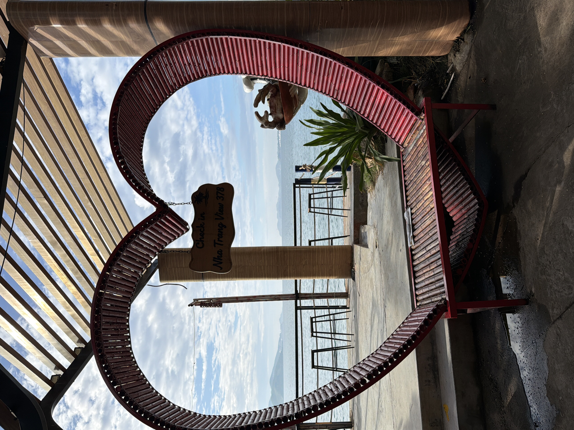 A large, red, heart-shaped bench with a slatted bamboo seat and back is positioned in the foreground, framing a view of the sea and sky. Above the bench, a wooden sign with text is suspended by a chain. The sign reads "Check in Nha Trang View 378". Beyond the bench and sign, a promenade runs along the coastline, with several high stools visible near the water's edge. A railing separates the promenade from the sea. In the distance, a chain of islands and the sea under a cloudy sky are visible. A large, wooden pillar with a wood grain texture stands to the right of the heart-shaped bench. The ground in front of the bench is wet and reflective, indicating recent rain or water.