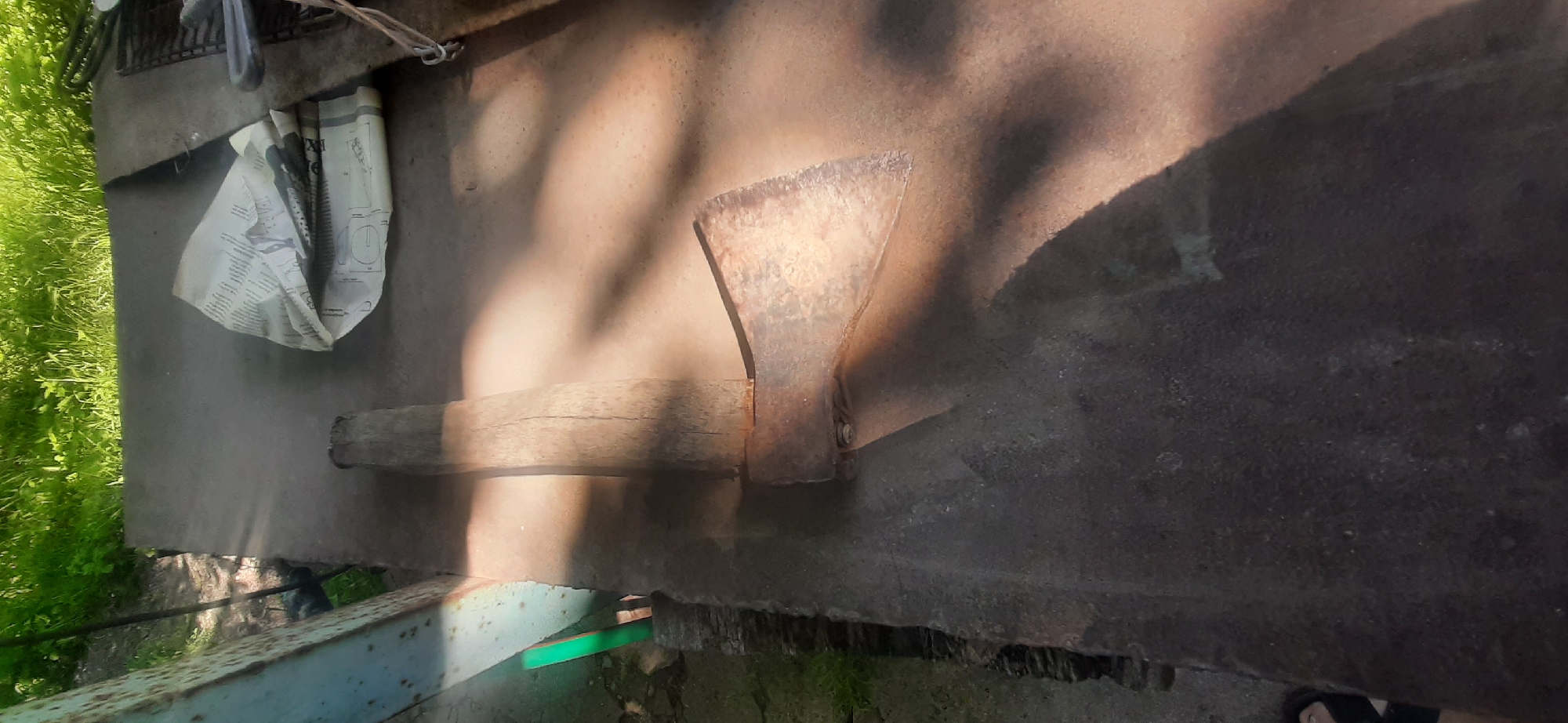 An axe with a wooden handle is lying on a dark, flat surface, possibly a workbench, outdoors. The axe head is made of rusted metal and is positioned diagonally across the surface. A crumpled piece of newspaper or paper is visible to the right of the axe. In the background, there is green grass and foliage, suggesting an outdoor setting. The lighting indicates a sunny day, with distinct shadows cast by the axe and other unseen objects. The overall scene appears to be in Yessentukskaya, Russia, given the context provided. No people are directly visible in the frame, although a partial view of a foot or shoe is present in the lower left corner.